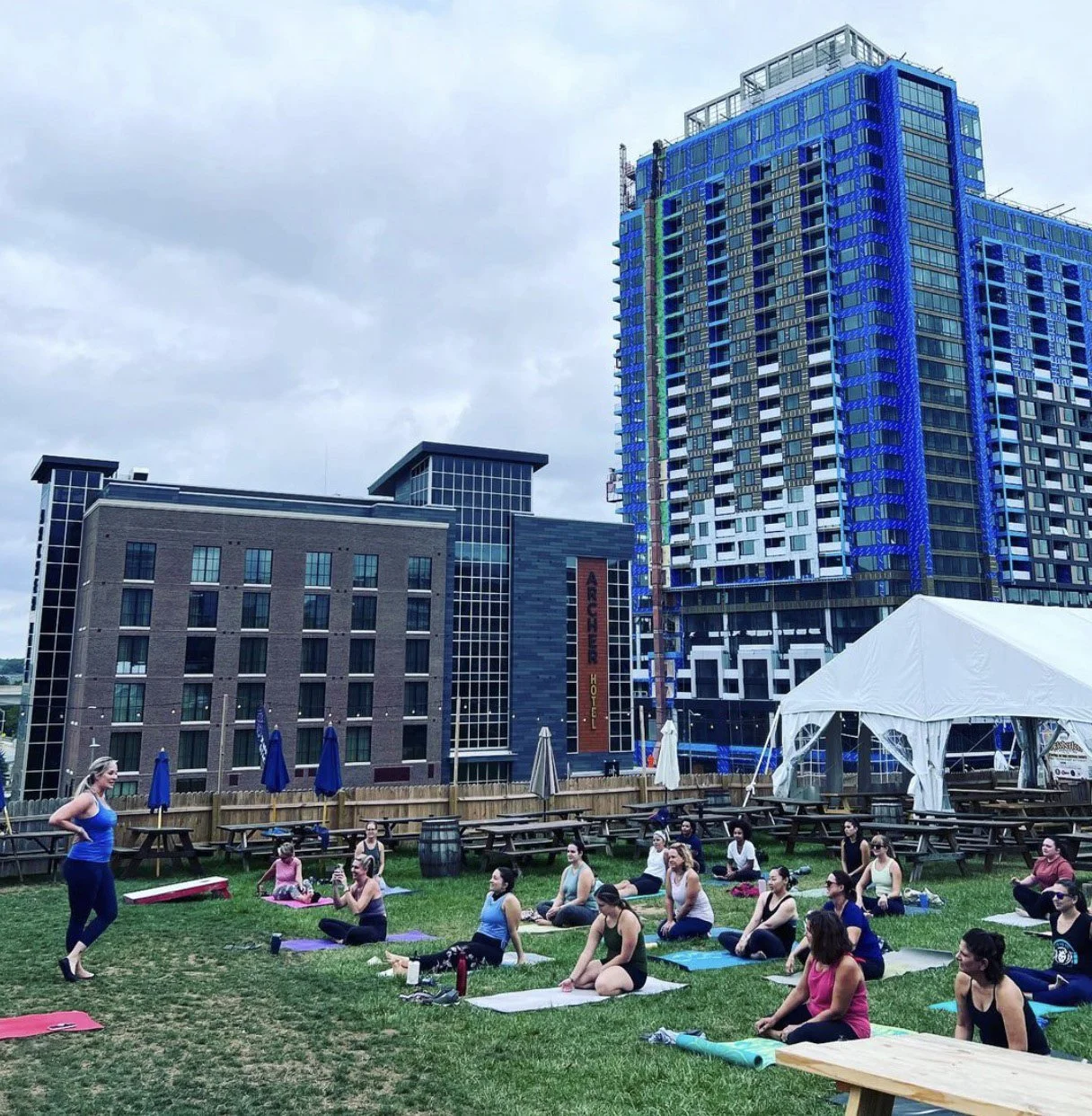 a group of people gather on the lawn at shipgarten on yoga mats, facing an instructor. The shipgarten picnic tables have been moved to the side. in the background, a tall new building is under construction