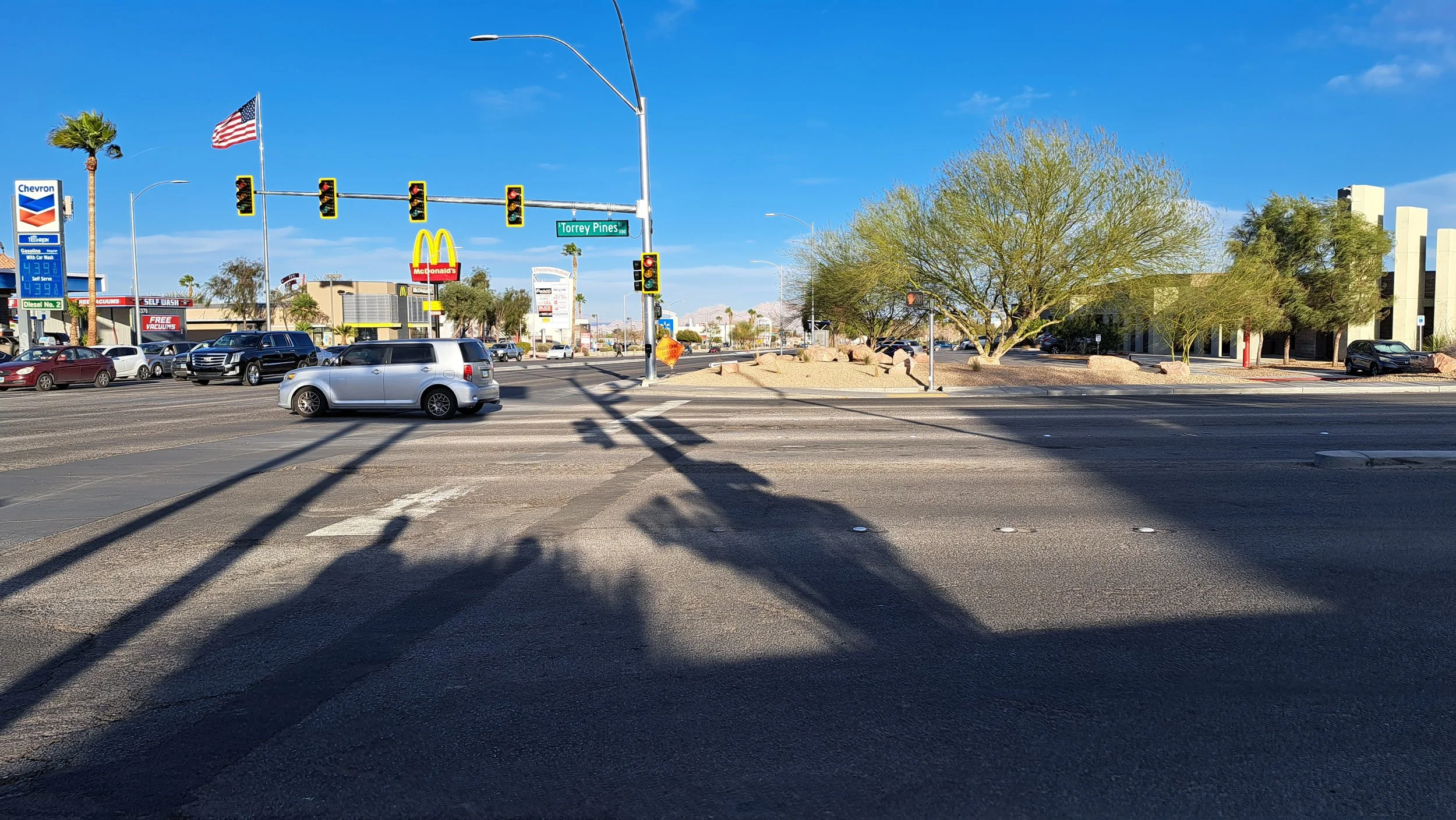 Street level photo of a large intersection in Charleston, with strip malls in the background andmultiple lanes of traffic in each direction, narrow sidewalks, and a faded crosswalk marking. There are several cars but no people walking