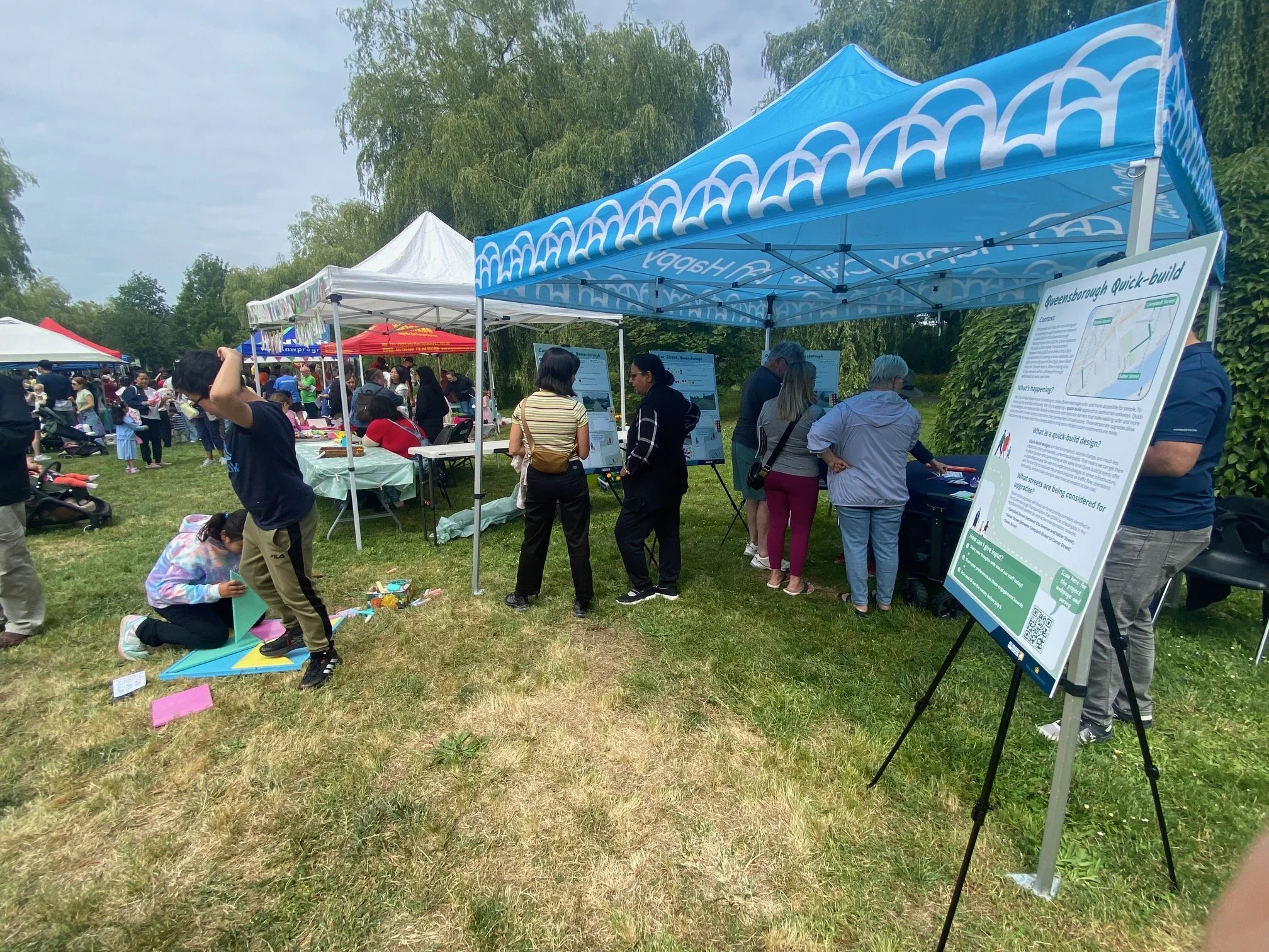 People gathered under and around a blue Happy Cities tent at an outdoors pop-up event.