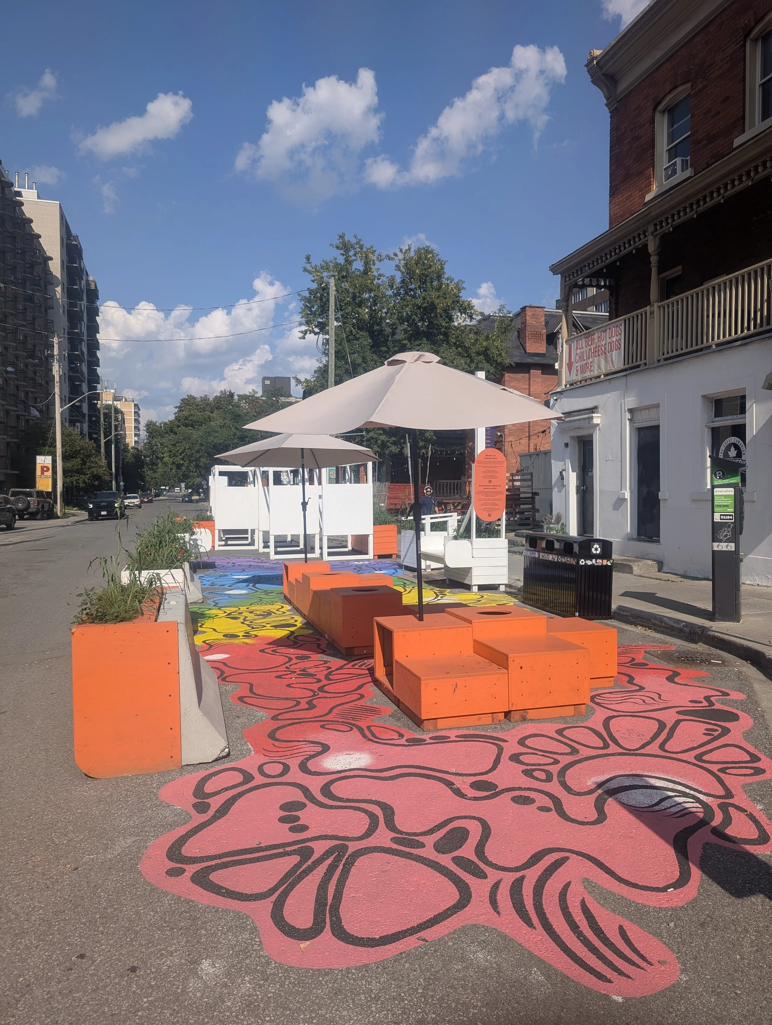 Photo of the McLaren st activation, with colourful wood boxes painted orange and doubling as seating under two shade umbrellas. Half the street is closed to make space for the plaza