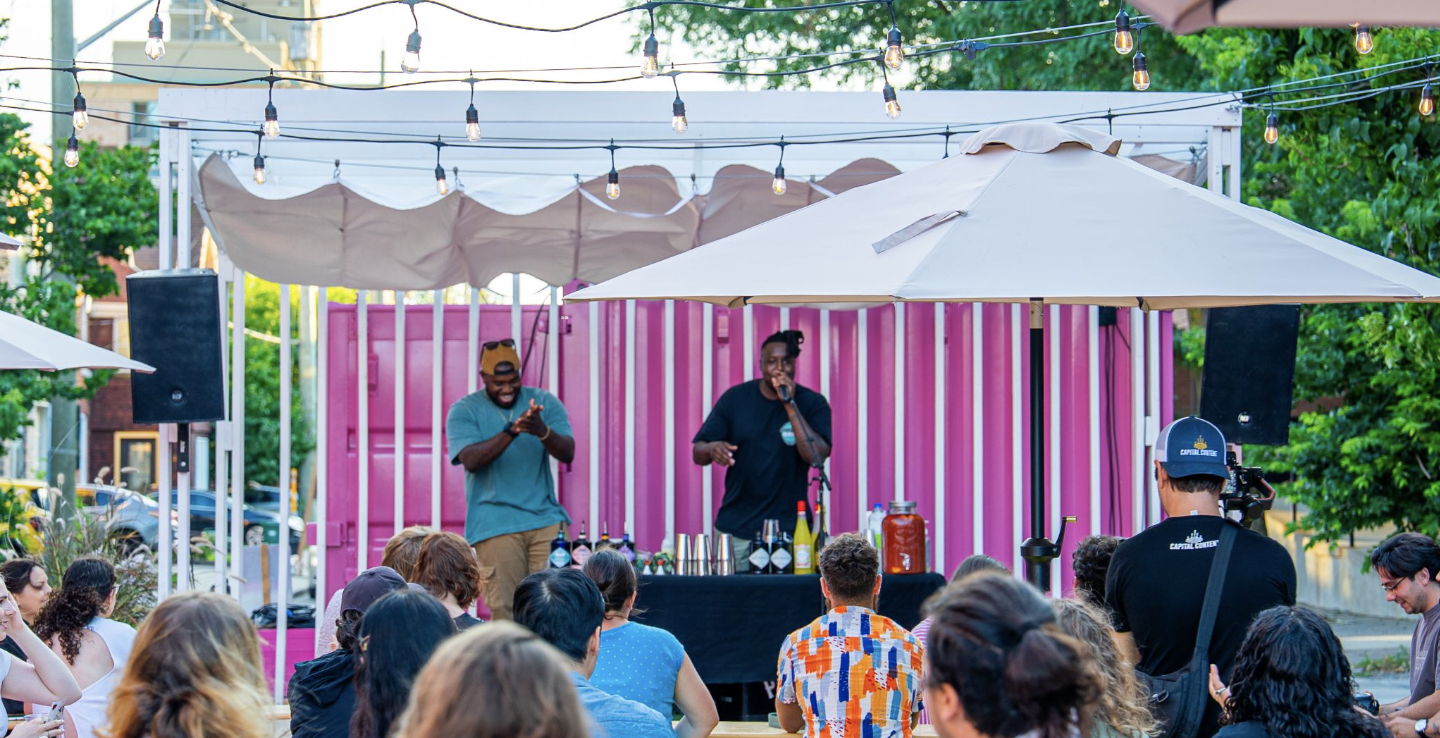 Photo of a temporary stage during the summer at a street plaza. Dozens of people sit in the plaza to watch a event put on by two performers