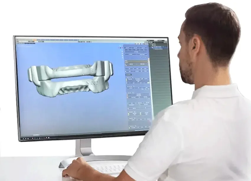 A dental technician at a computer workstation, reviewing a 3D CAD model of a custom-fit oral appliance used for snoring and sleep apnoea therapy.