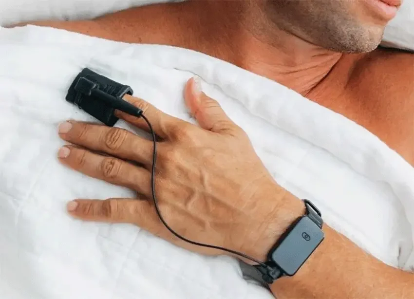 A close-up of a person in bed using a finger pulse oximeter and wrist monitor, which are used in home sleep studies to diagnose sleep apnoea.