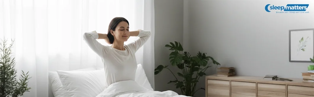 Woman sits up and stretches in bed in a bright bedroom, showing improved sleep quality that people often seek when managing sleep apnoea.
