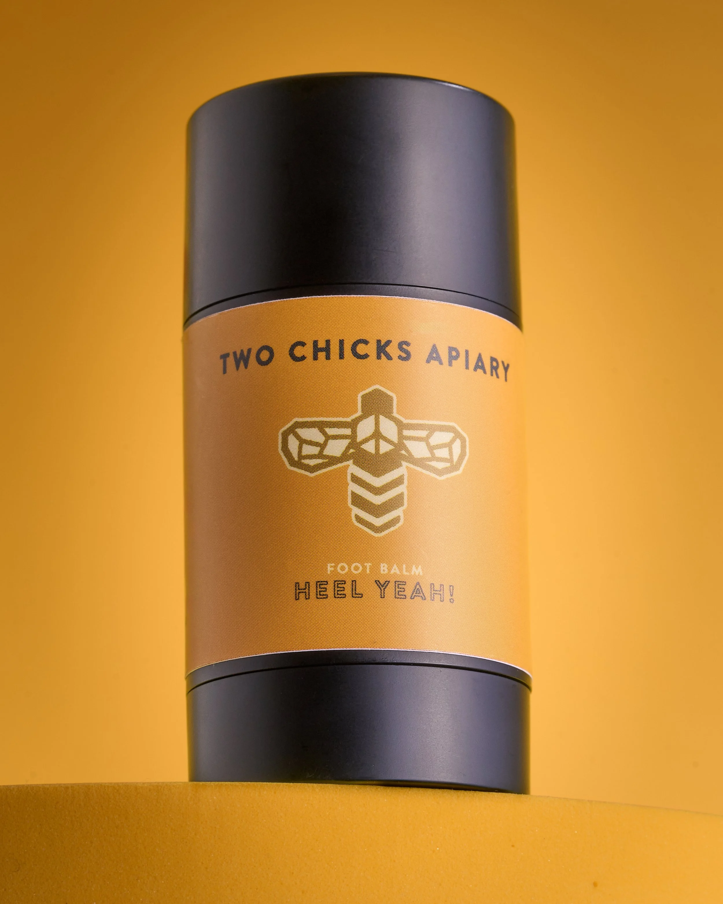 A small black cylindrical container of foot balm labeled 'Two Chicks Apiary Foot Balm Heel Yeah!' placed on a wooden surface.
