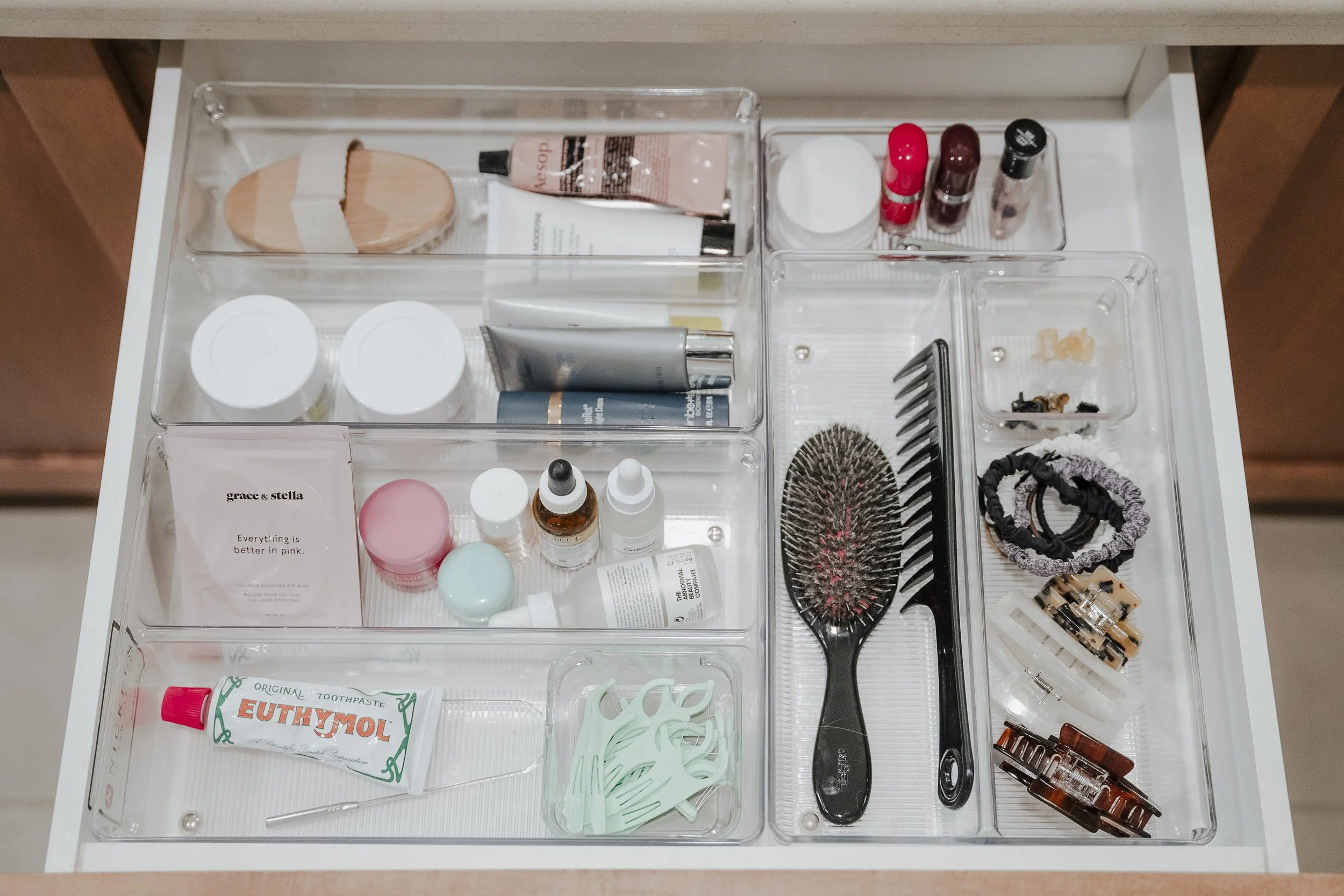 Open drawer containing hair and beauty accessories, including hairbrushes, clips, elastics, hair ties, skincare products, toothpaste, and small containers.