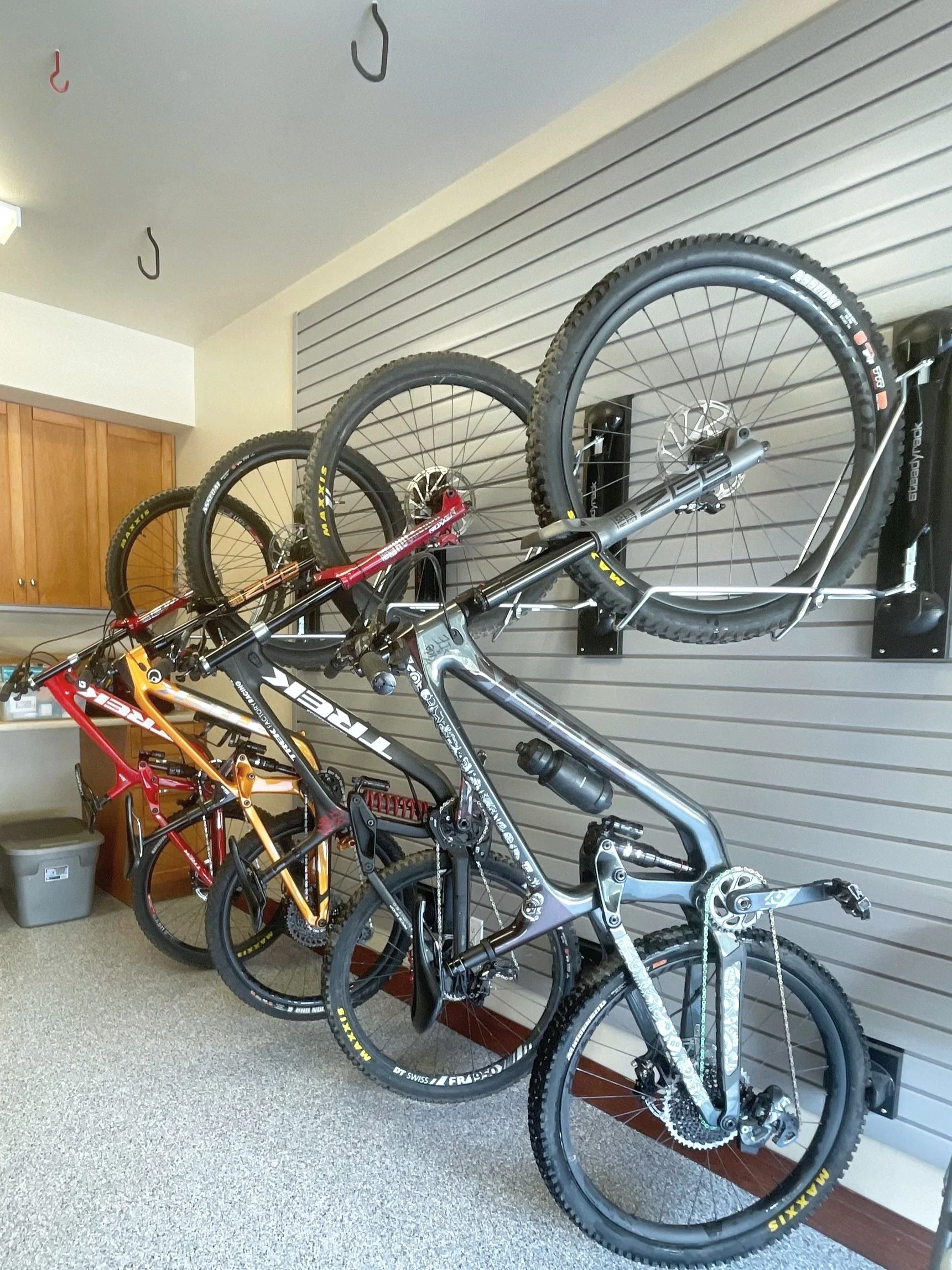 A metal carbon fiber mountain bike with front suspension, mounted on a striped grey wall, with three other bikes in red, yellow, and black colors, stored inside with wheels removed and hung on hooks.