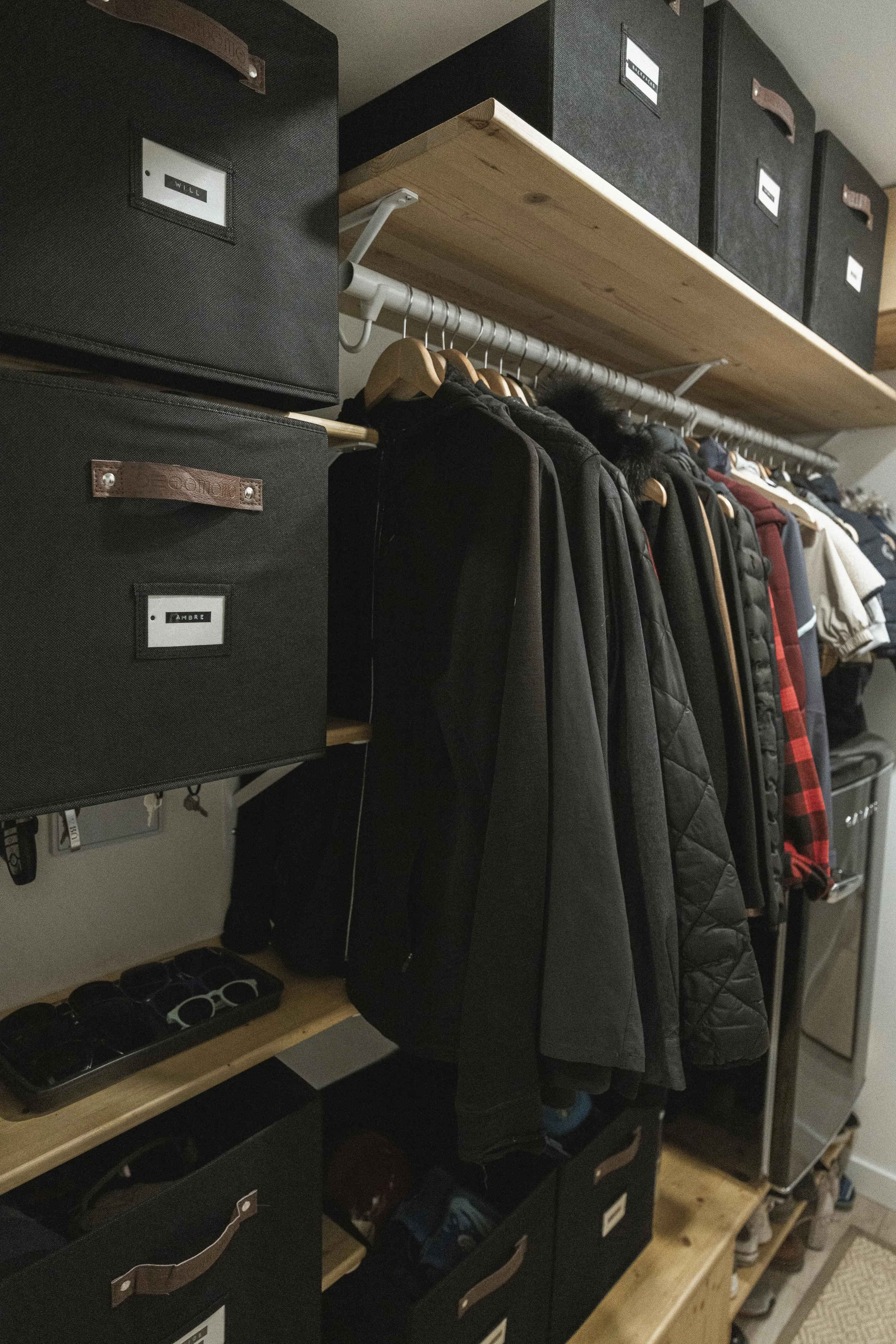 An organized closet with black storage boxes, a wooden shelf, and coats hanging on a metal rod.