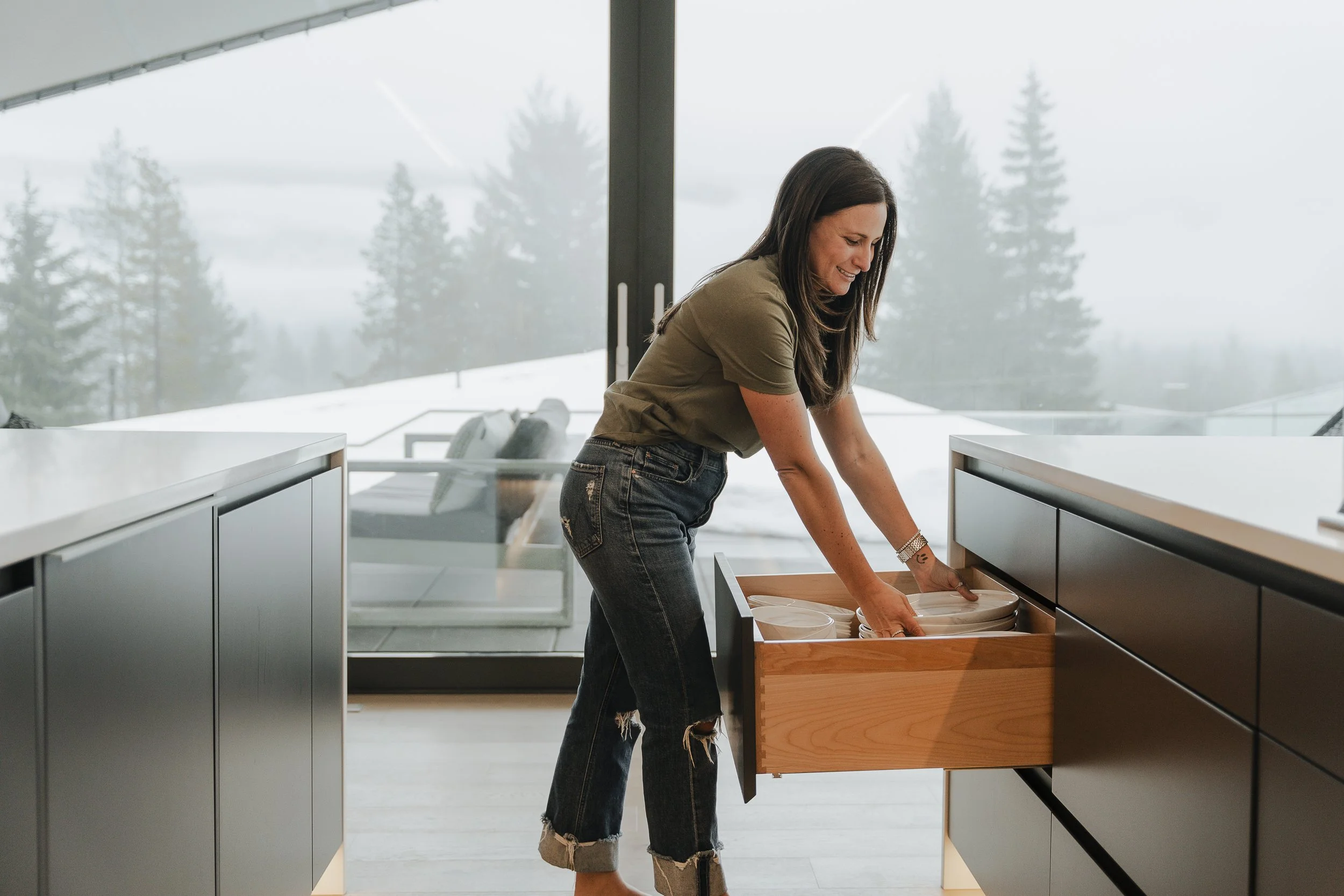 Woman opening a drawer in a modern kitchen with a snowy mountain view through large windows in the background.