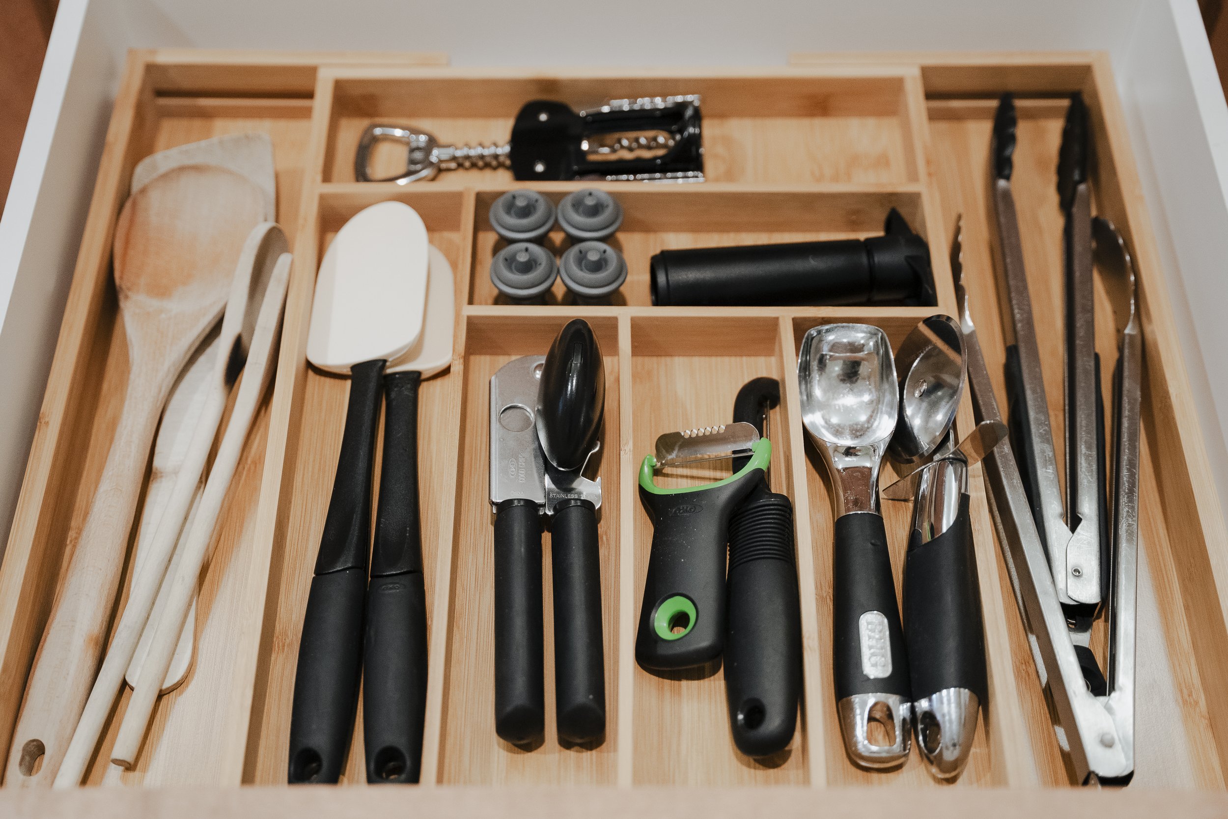 Kitchen utensil drawer containing spoons, spatulas, tongs, can opener, vegetable peeler, garlic press, and other utensils organized in compartments.