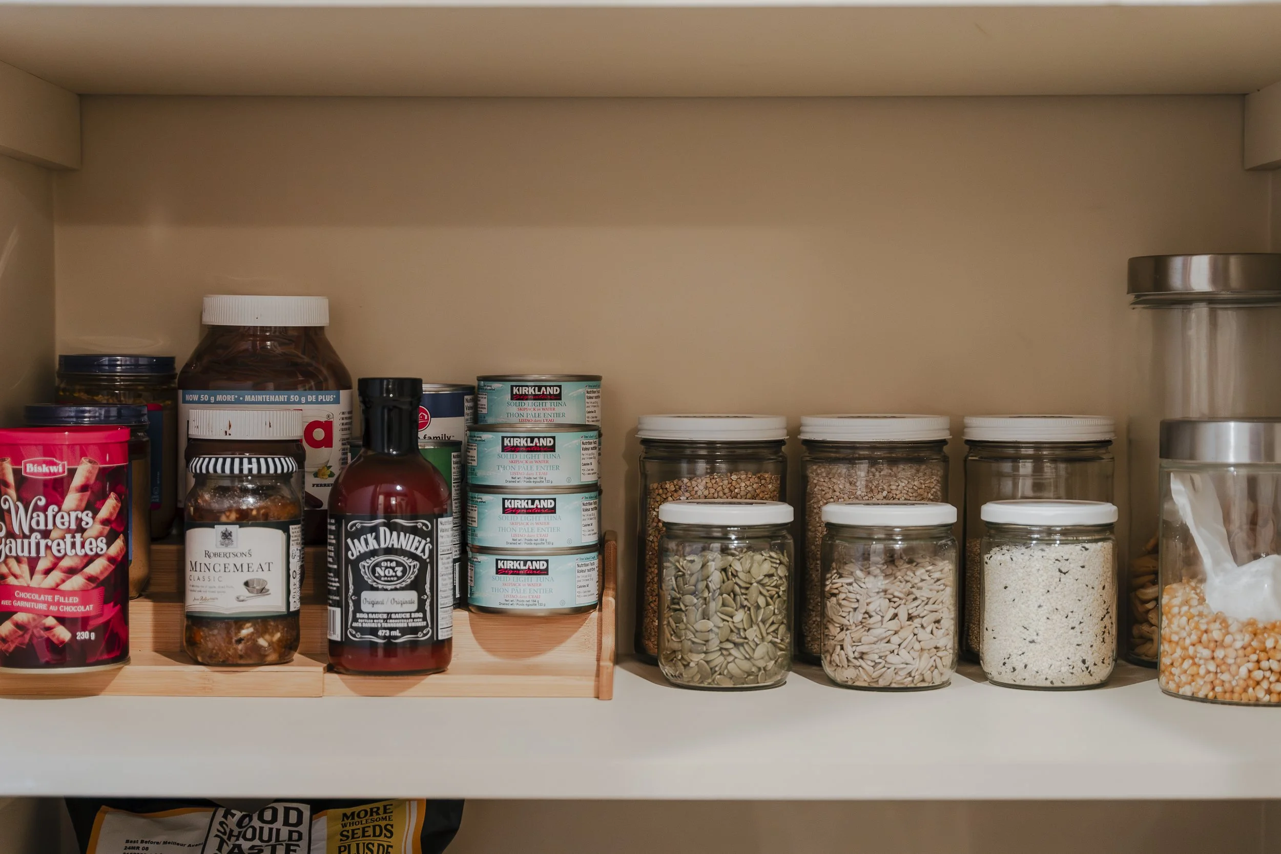 Pantry shelf containing jars of seeds, canned tuna, spices, and snack items.