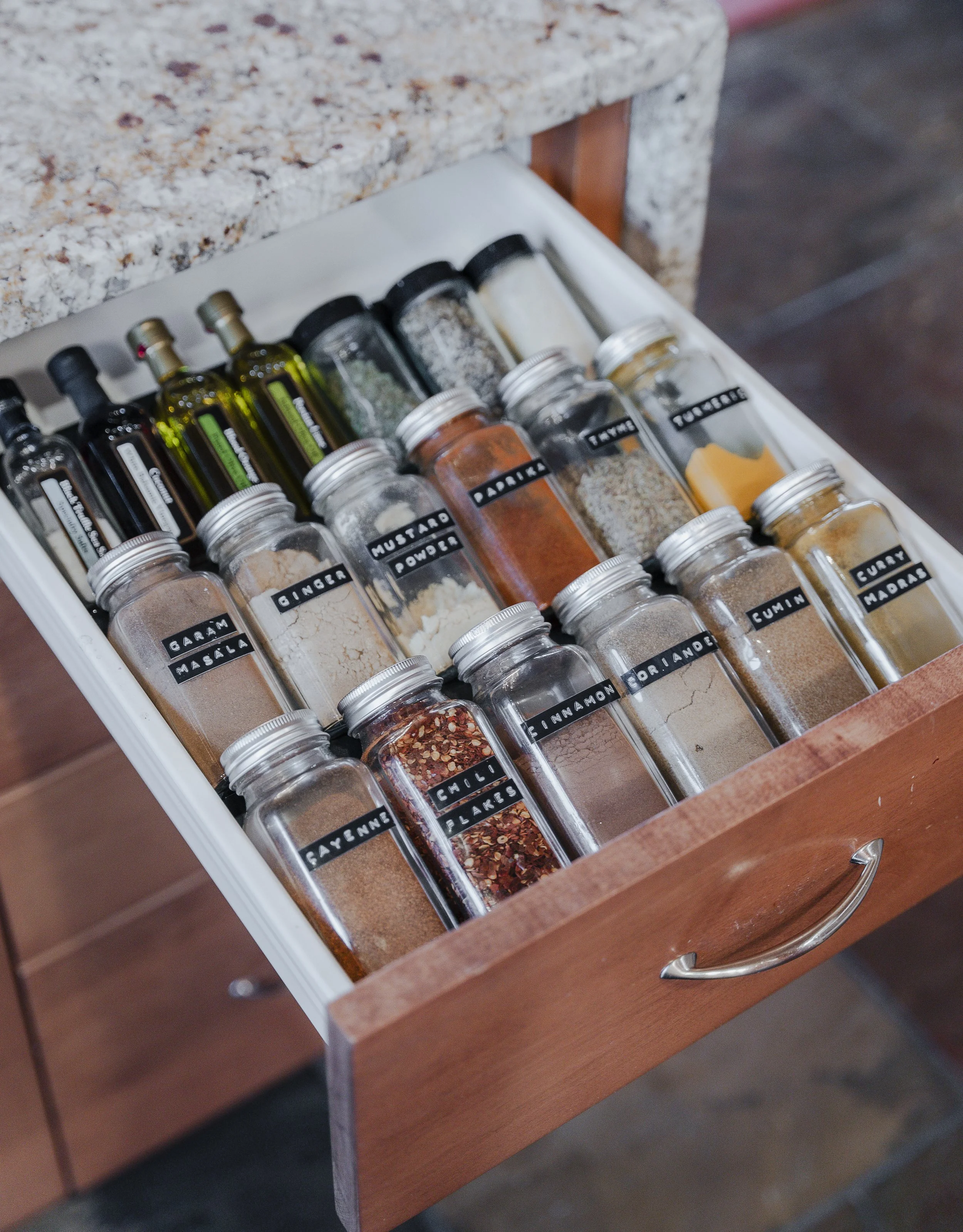 An open drawer containing glass jars and bottles filled with various spices and herbs, with labels such as cumin, turmeric, cayenne, garlic, and paprika.