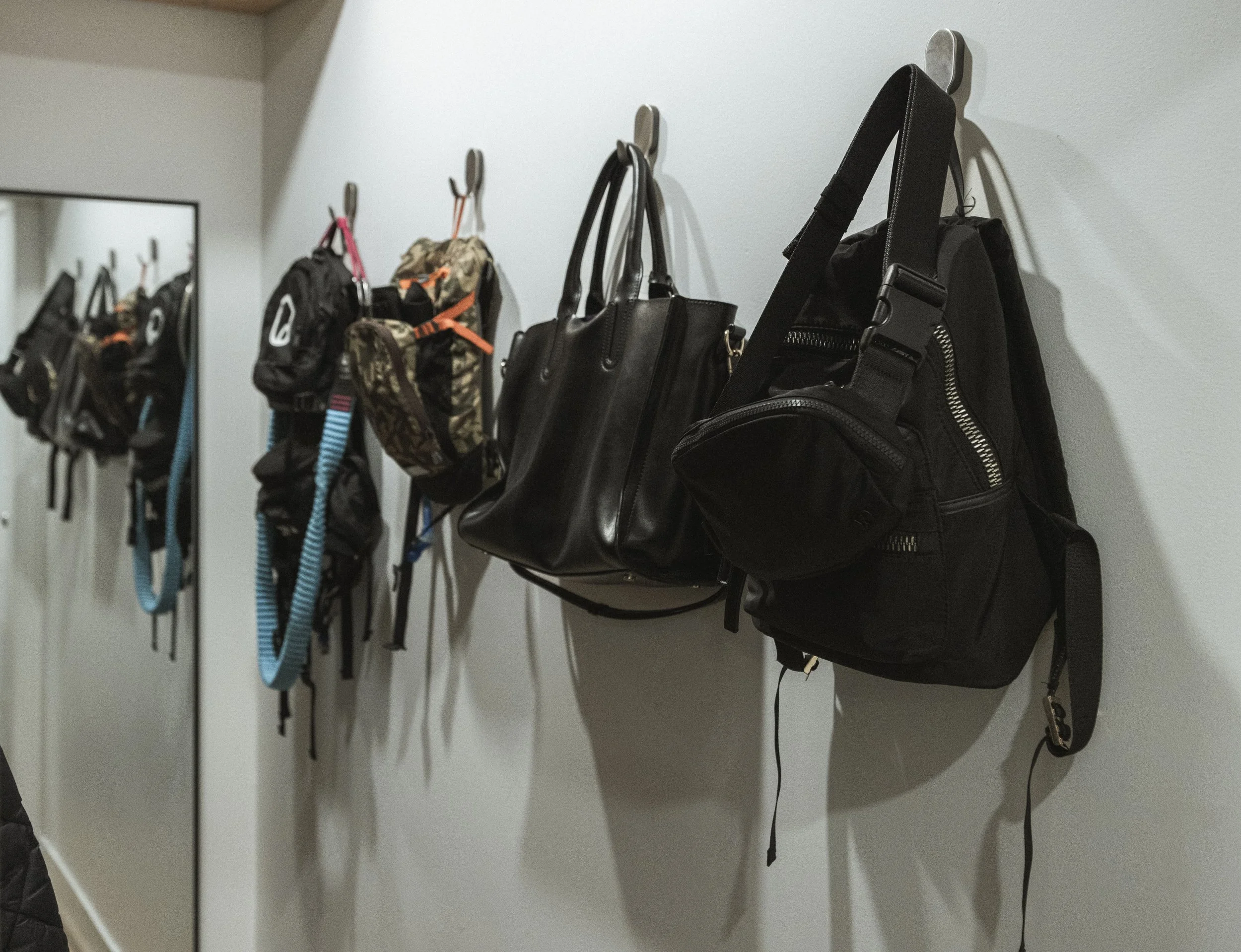 Wall-mounted hooks holding several bags and backpacks near a mirror.