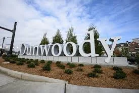Large white Dunwoody sign with greenery in the background under a blue sky.