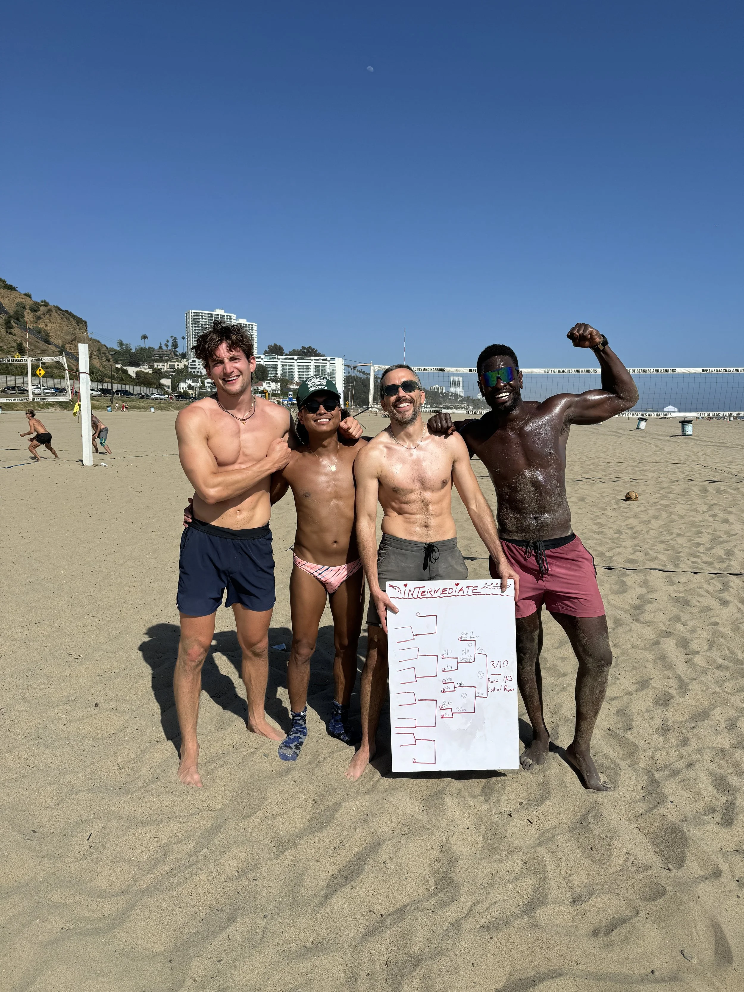 Four shirtless men standing on a sandy beach, smiling and holding a tournament bracket board, with a volleyball net and other beachgoers in the background.