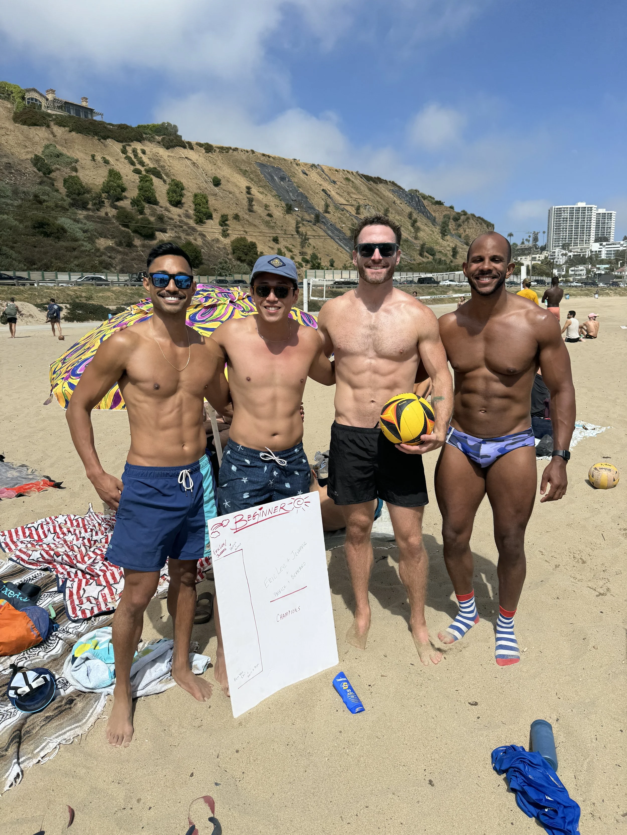 Four men in swimwear standing on a beach, smiling, with a hill, buildings, and other beachgoers in the background.