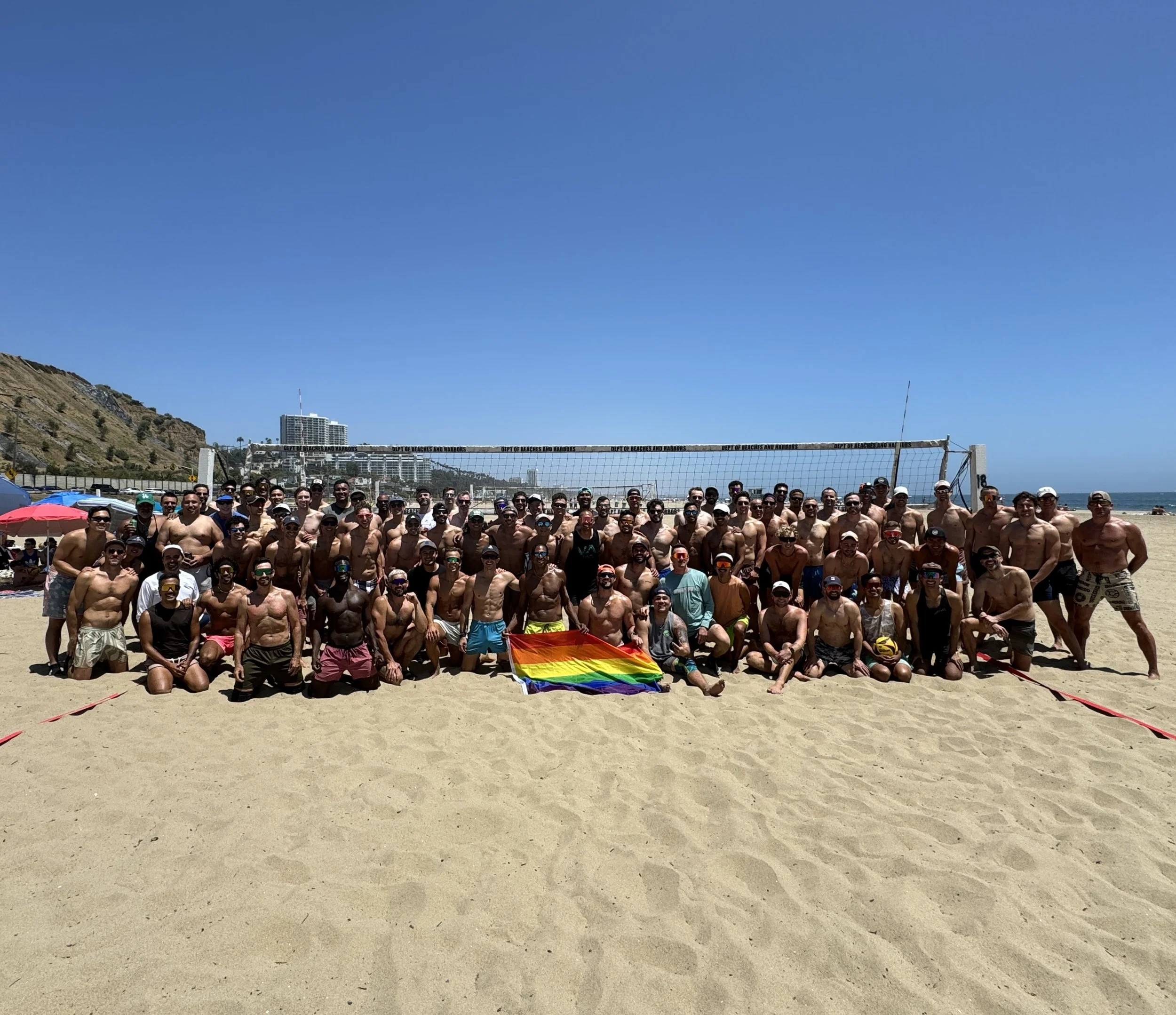 Group of shirtless men on a beach holding a rainbow pride flag, with a volleyball net and ocean in the background.