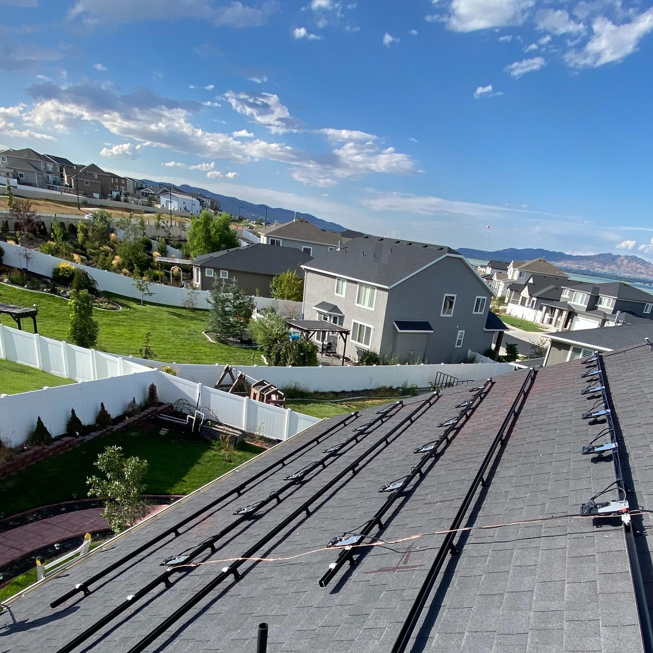 Residential neighborhood with houses, front yards, and solar panel mounting racks on a roof under a clear blue sky.