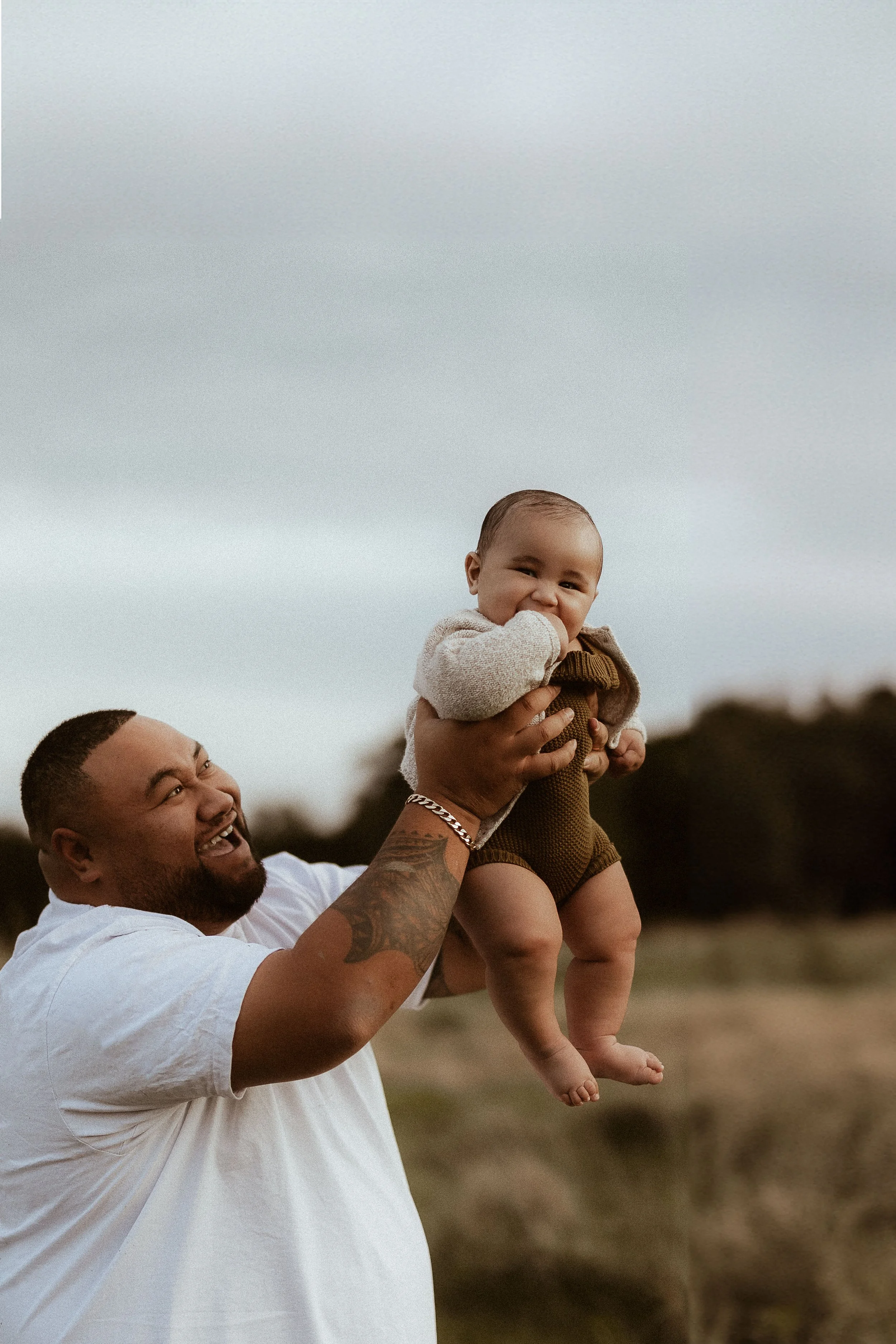 A man with a beard and tattoos on his arm holding a happy baby, laughing and playing outdoors on a cloudy day.