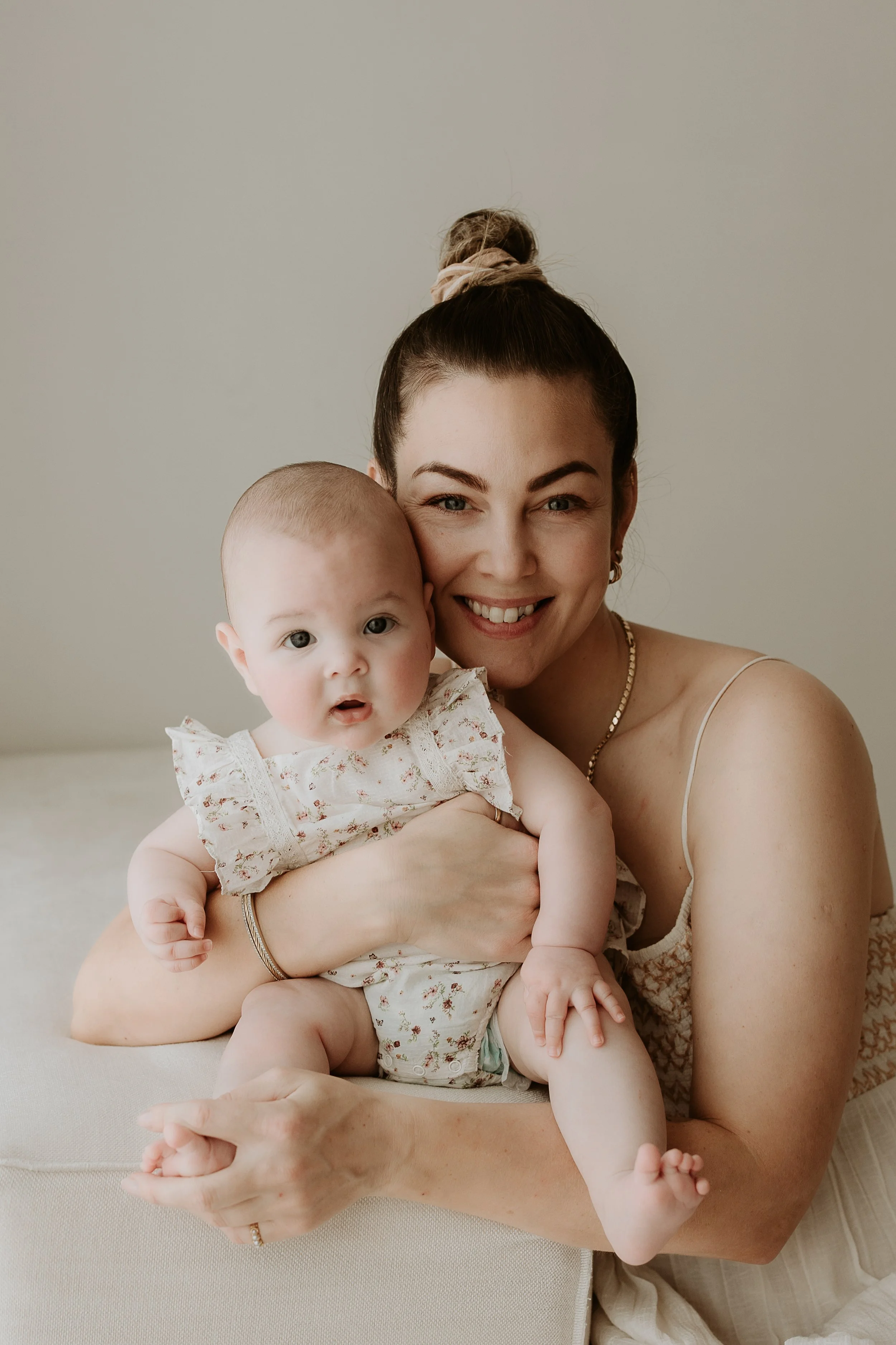 A woman smiling and holding a baby, both looking at the camera, sitting indoors with a neutral background