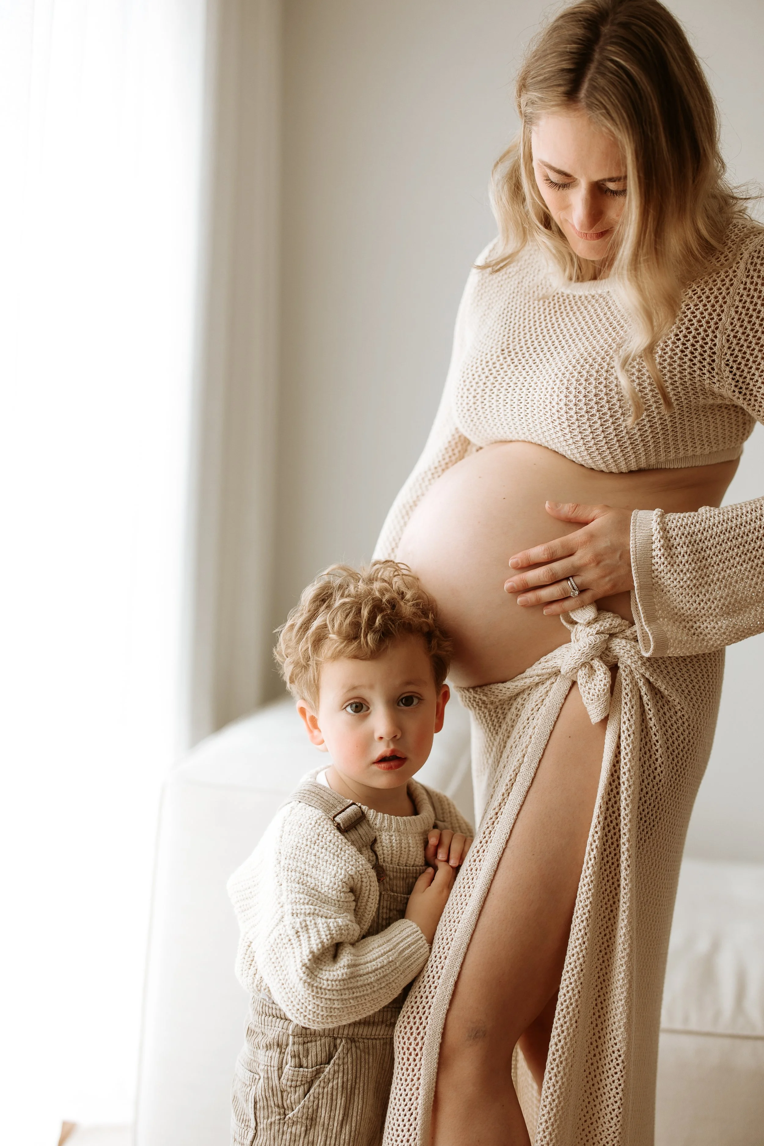 Pregnant woman in beige outfit with her young son, who is wearing a beige sweater, standing in a bright room