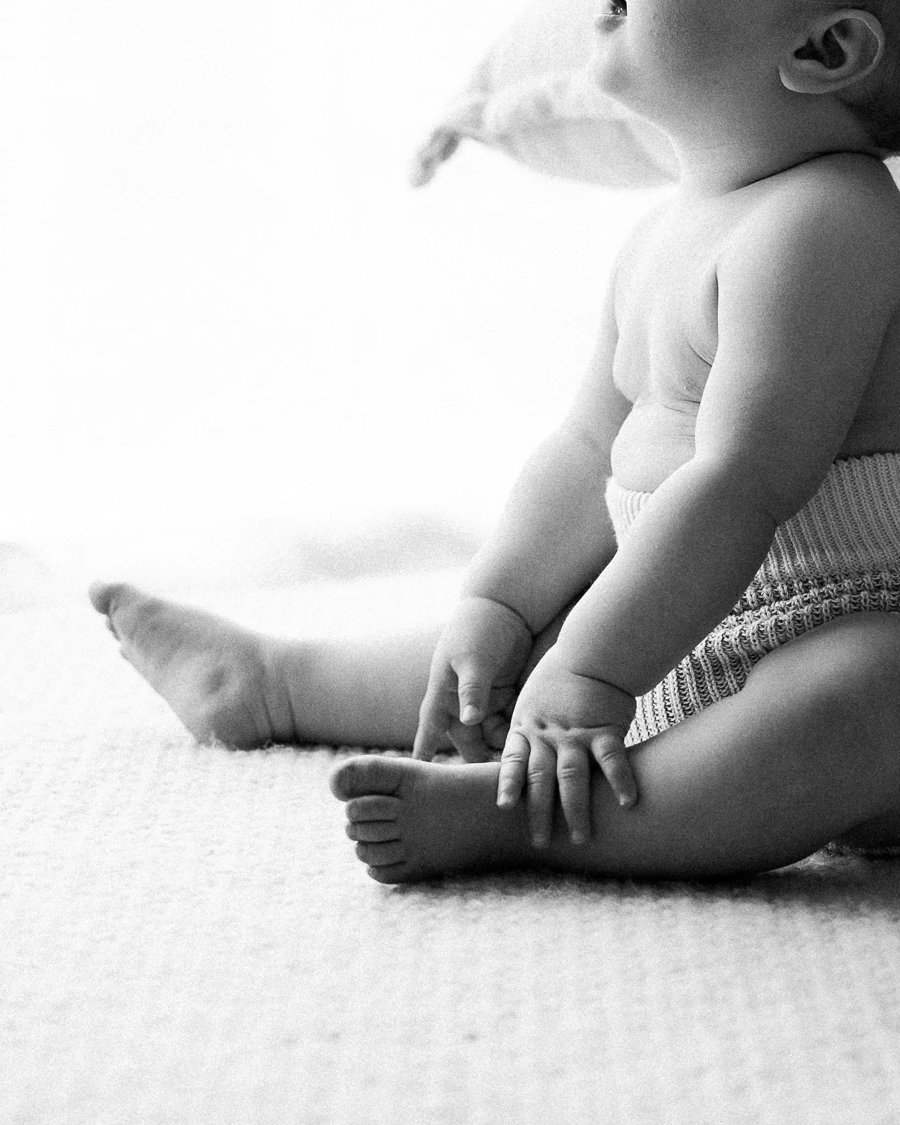 Black and white photo of a young child sitting on the floor, touching their toes with one hand, wearing a diaper and striped shorts, with a soft blanket in the background.