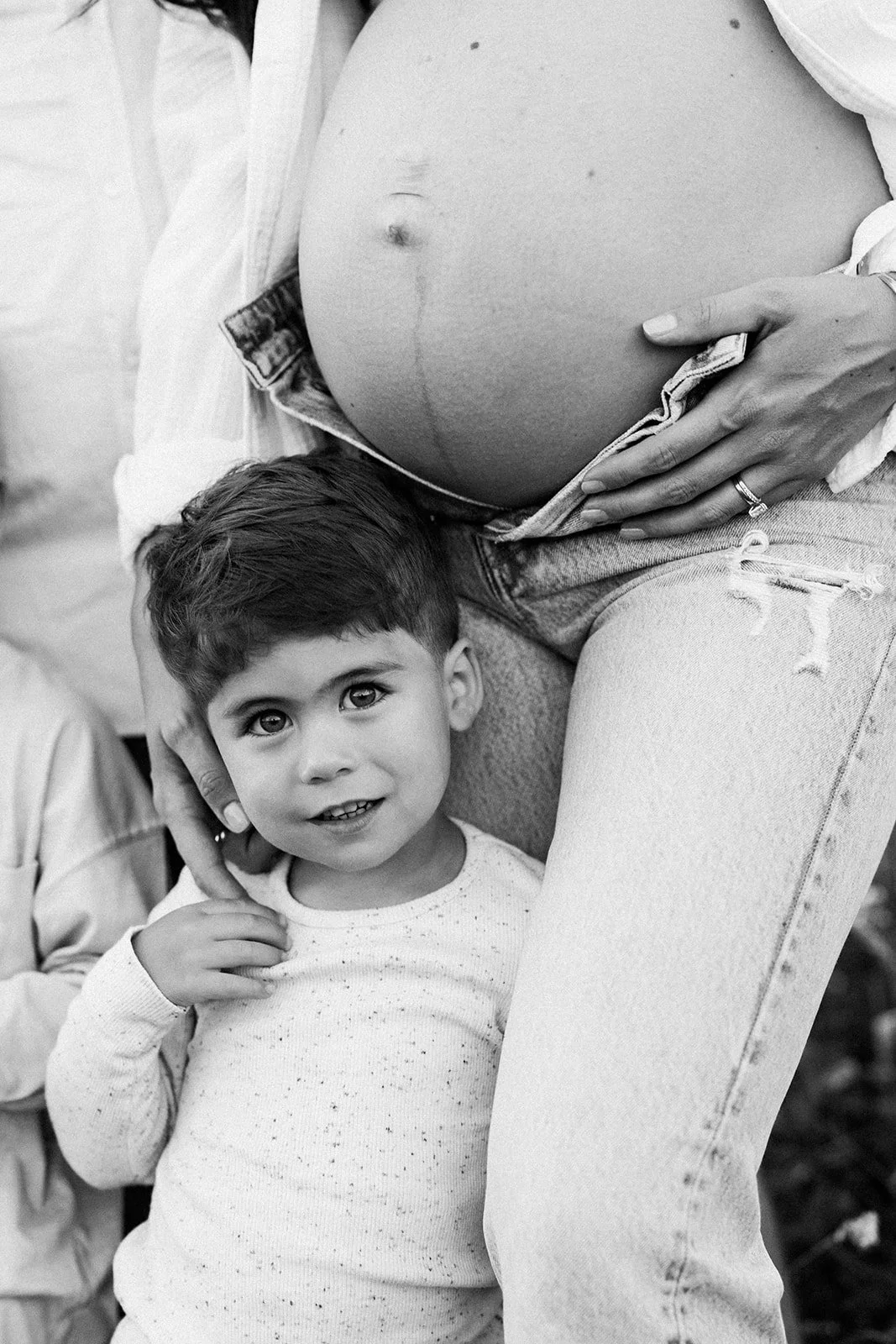 A pregnant woman with her hand on her belly and a young boy standing beside her, touching her leg, looking at the camera, in black and white.