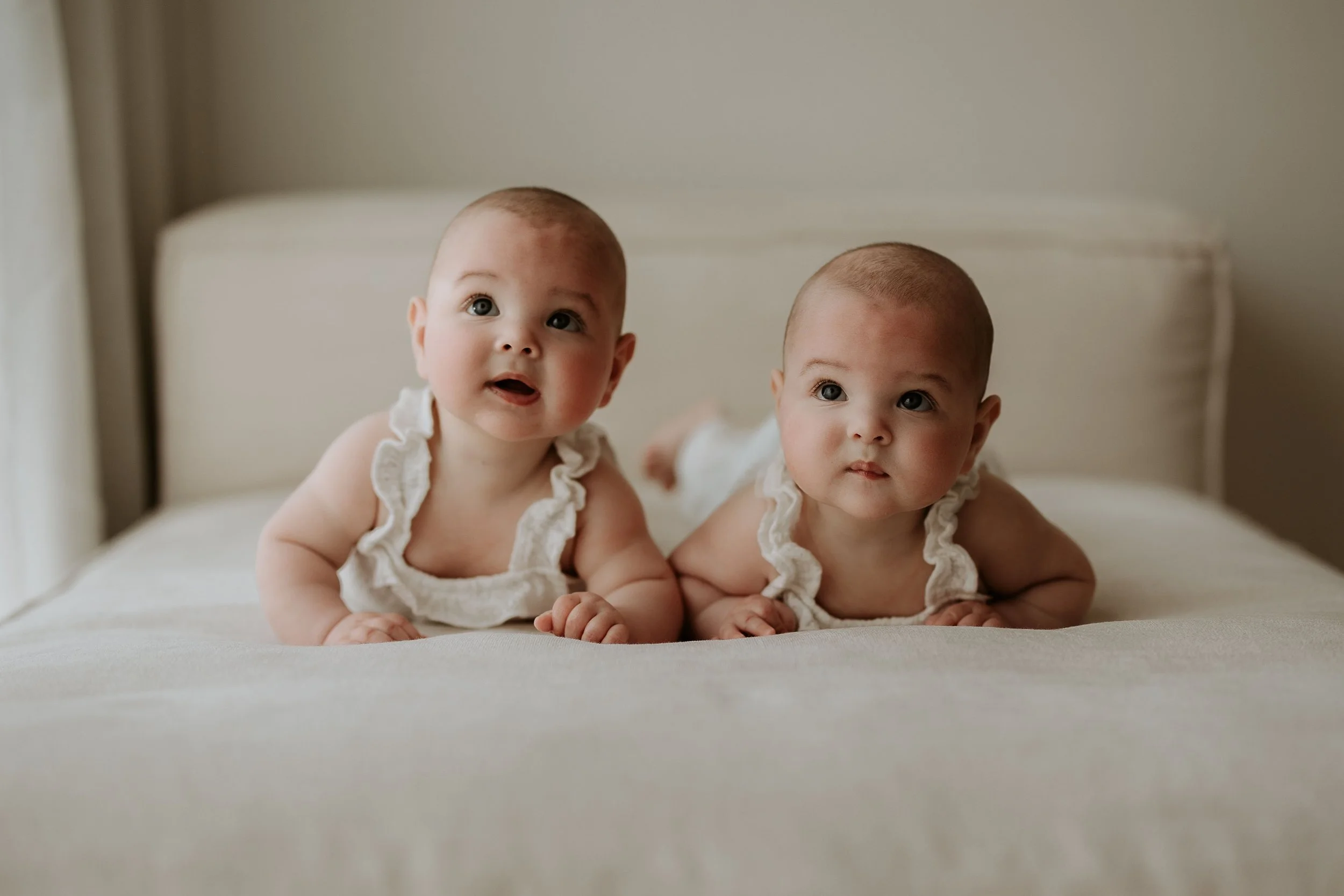 Two babies lying on their stomachs on a bed, looking up. Both babies are wearing white ruffled sleeveless outfits and have short light brown hair. The background shows a cream-colored headboard and beige wall.