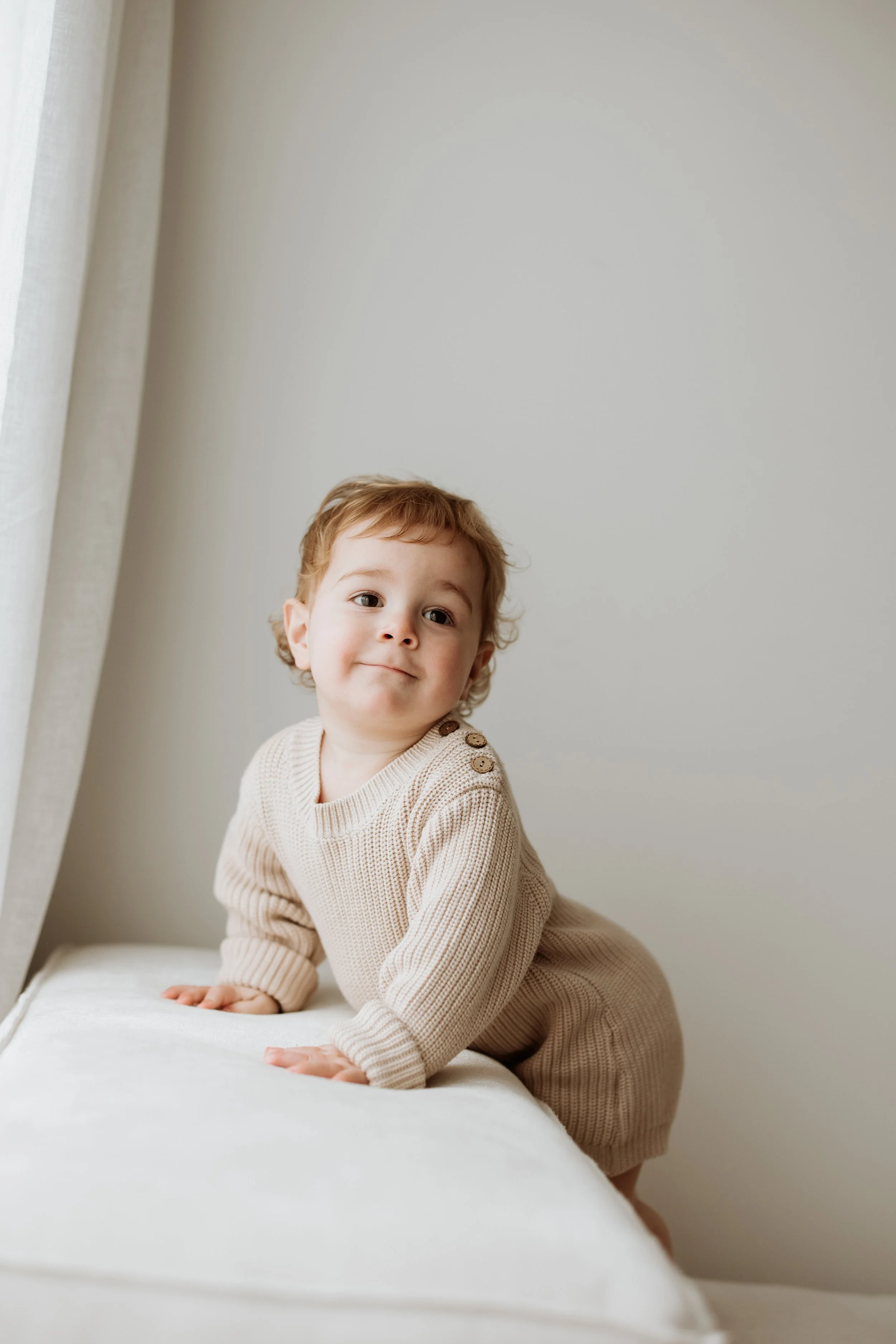 A toddler with light brown curly hair and wearing a beige knit outfit with buttons on the shoulder, leaning on a white surface near a window with light-colored curtains.