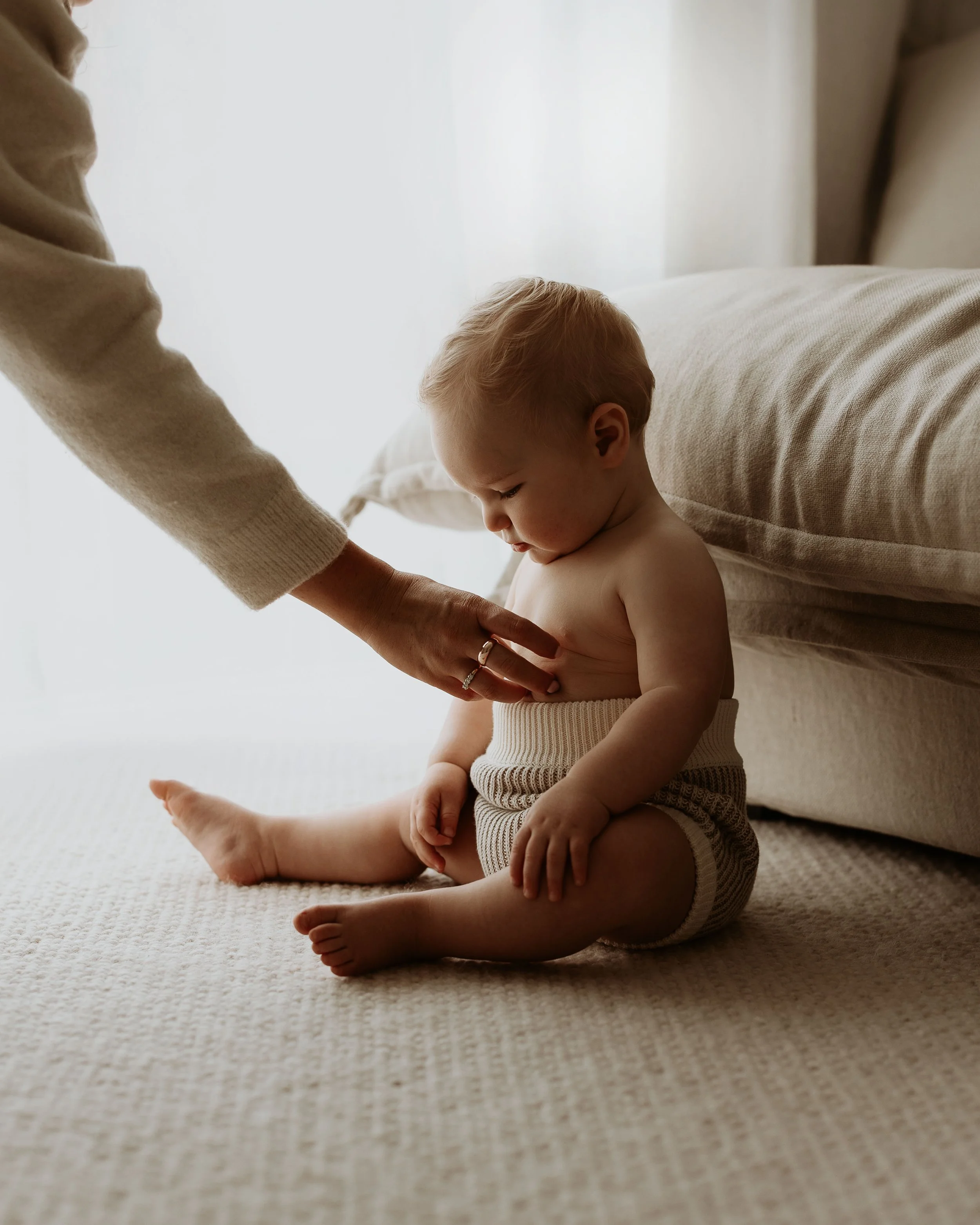 A young child sitting on a beige carpeted floor, looking down as an adult gently touches their stomach. The child is shirtless and wearing striped shorts, with a neutral indoor background.