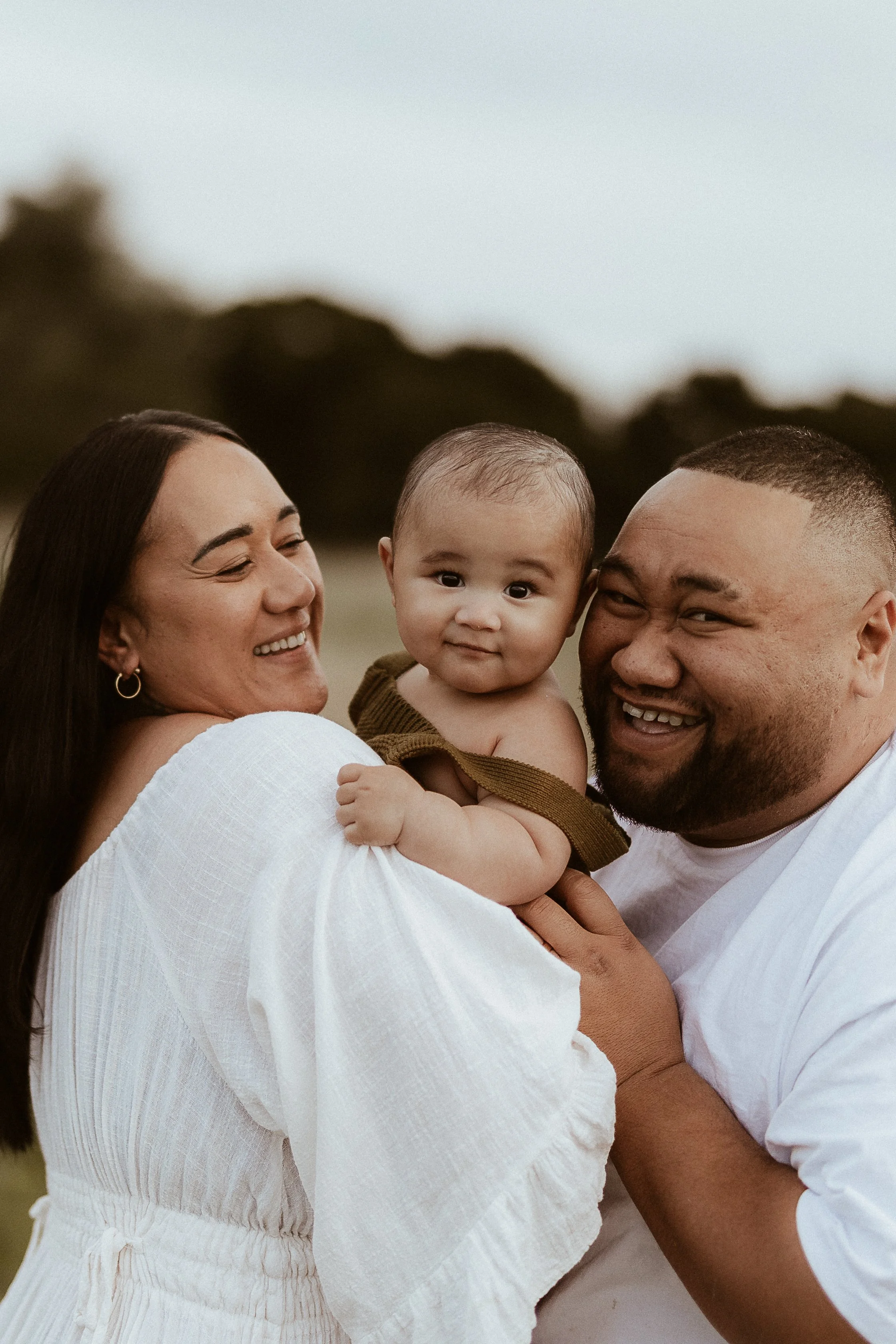 A happy family of three enjoying outdoor time, the mother holding the baby, the father smiling, with trees and sky in the background.