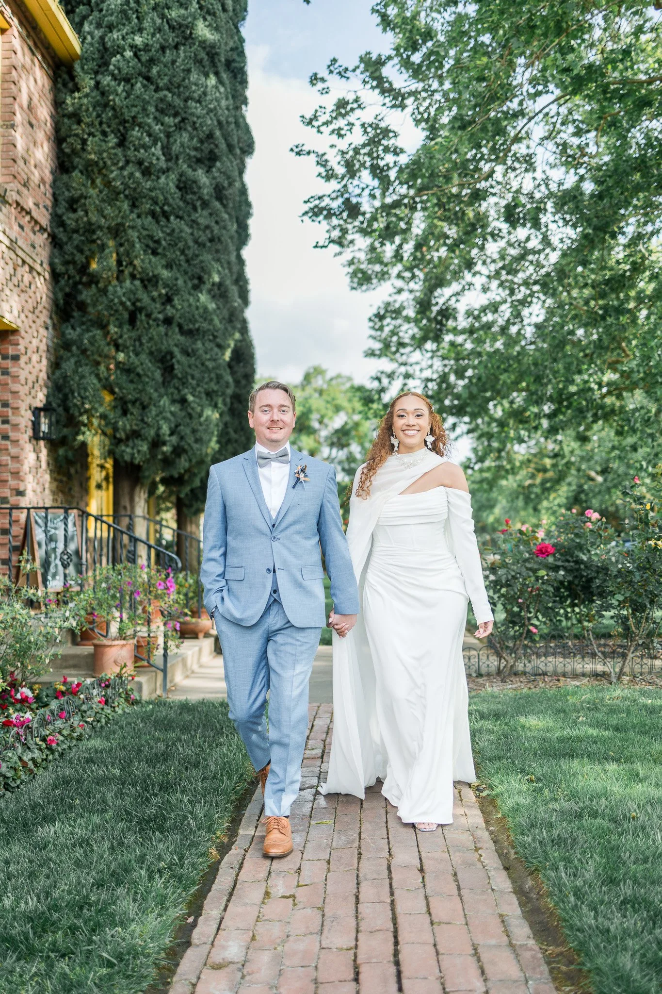 A newlywed couple walking hand-in-hand on a brick pathway outdoors, surrounded by greenery and flowers, with the bride in a white gown and the groom in a light blue suit.