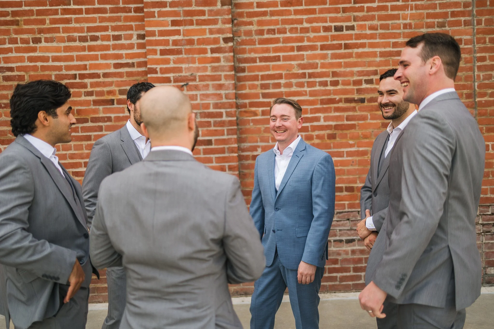 A group of six men in suits standing outside in front of a brick wall, engaging in a conversation and smiling.