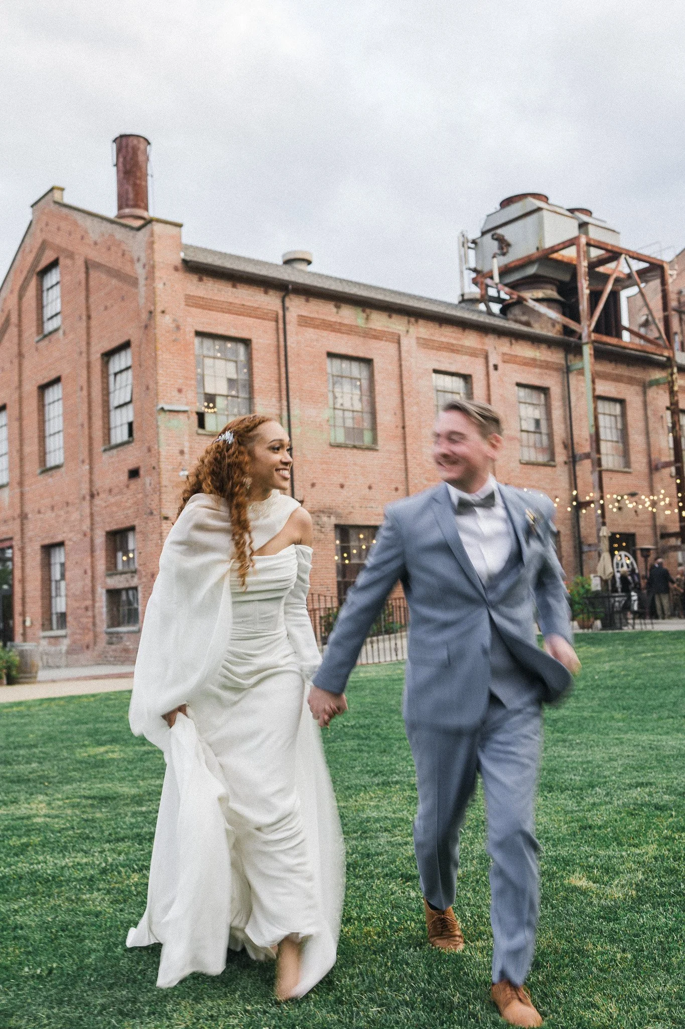 A bride and groom holding hands and smiling while walking on a grassy area outdoors, with a brick industrial building in the background.