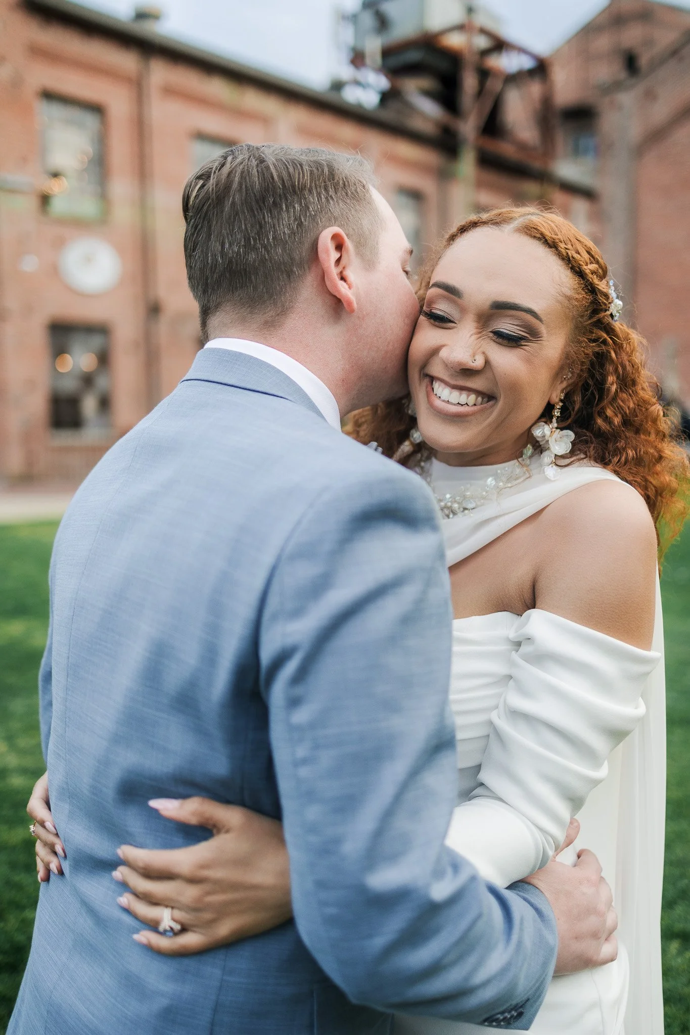 A smiling bride and groom sharing an intimate moment outdoors on a grassy area, with the groom kissing the bride's cheek. The bride has curly red hair and is wearing statement earrings and a white off-the-shoulder wedding dress. The groom is wearing 