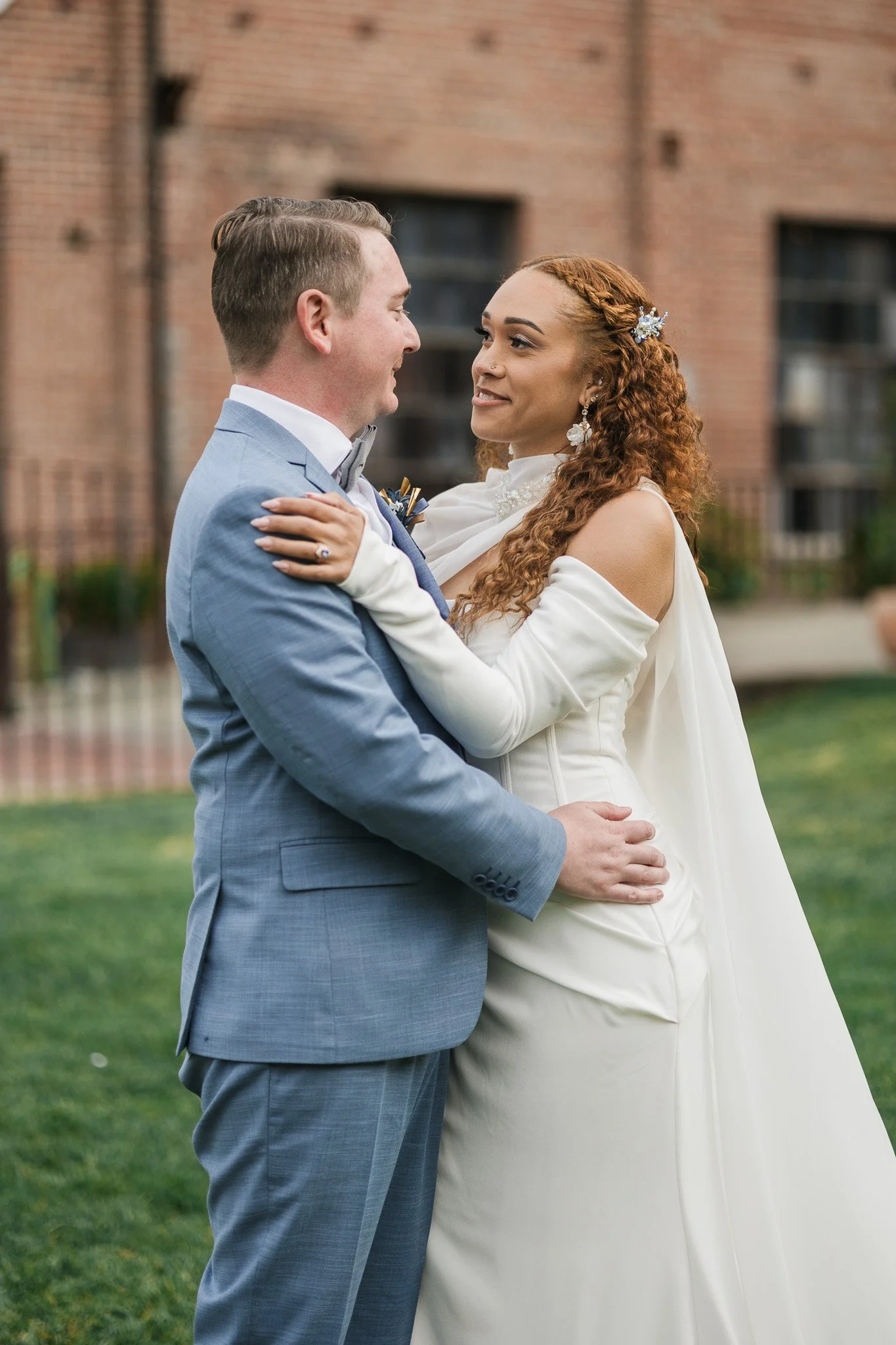 A bride and groom share a dance outdoors, with the bride wearing a white off-shoulder wedding gown and curly red hair, and the groom in a light blue suit and gray bow tie, in front of a brick building.