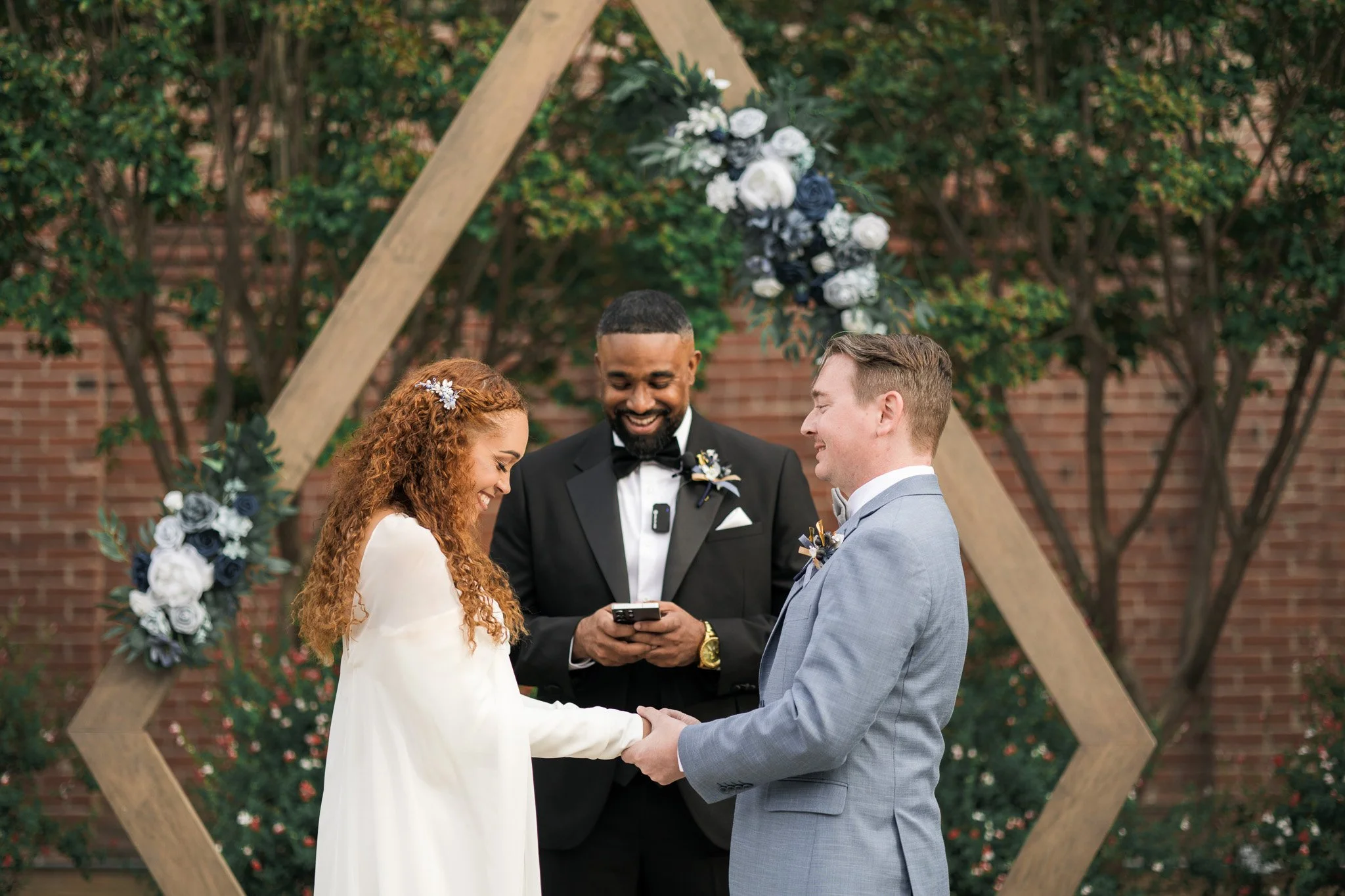 A couple getting married exchanging vows outdoors, with an officiant behind them, in front of a decorated wooden arch with flowers and greenery.
