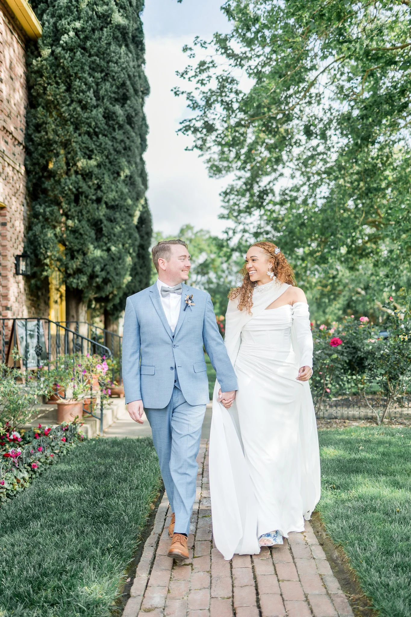 A newlywed couple walking hand-in-hand on a brick path in a garden, both smiling and looking at each other. The groom is wearing a light gray suit with a bow tie, and the bride is in a white wedding dress with off-the-shoulder sleeves and floral earr