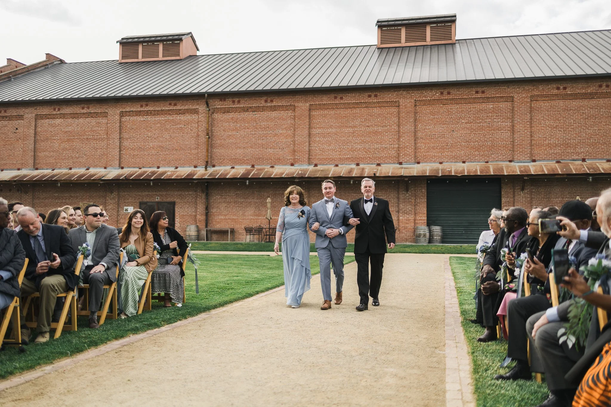 A wedding ceremony outdoors with guests seated on either side of a dirt aisle. A bride in a light blue dress walks arm-in-arm with two men in suits, one in black and the other in gray. The background features a large brick building and cloudy sky.