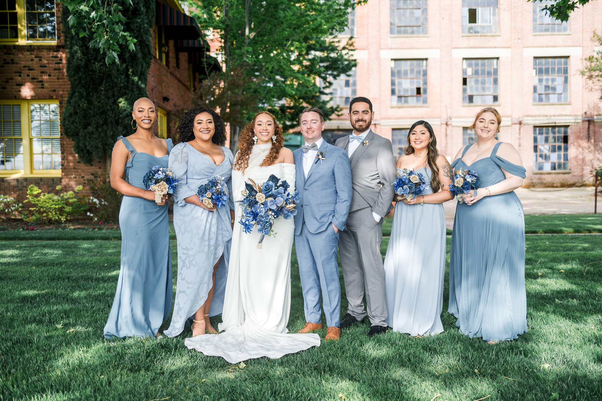 A wedding group photo featuring seven people, including the bride in a white dress and six bridesmaids and groomsmen in blue and gray, standing outdoors on a grassy area with trees and a brick building in the background.