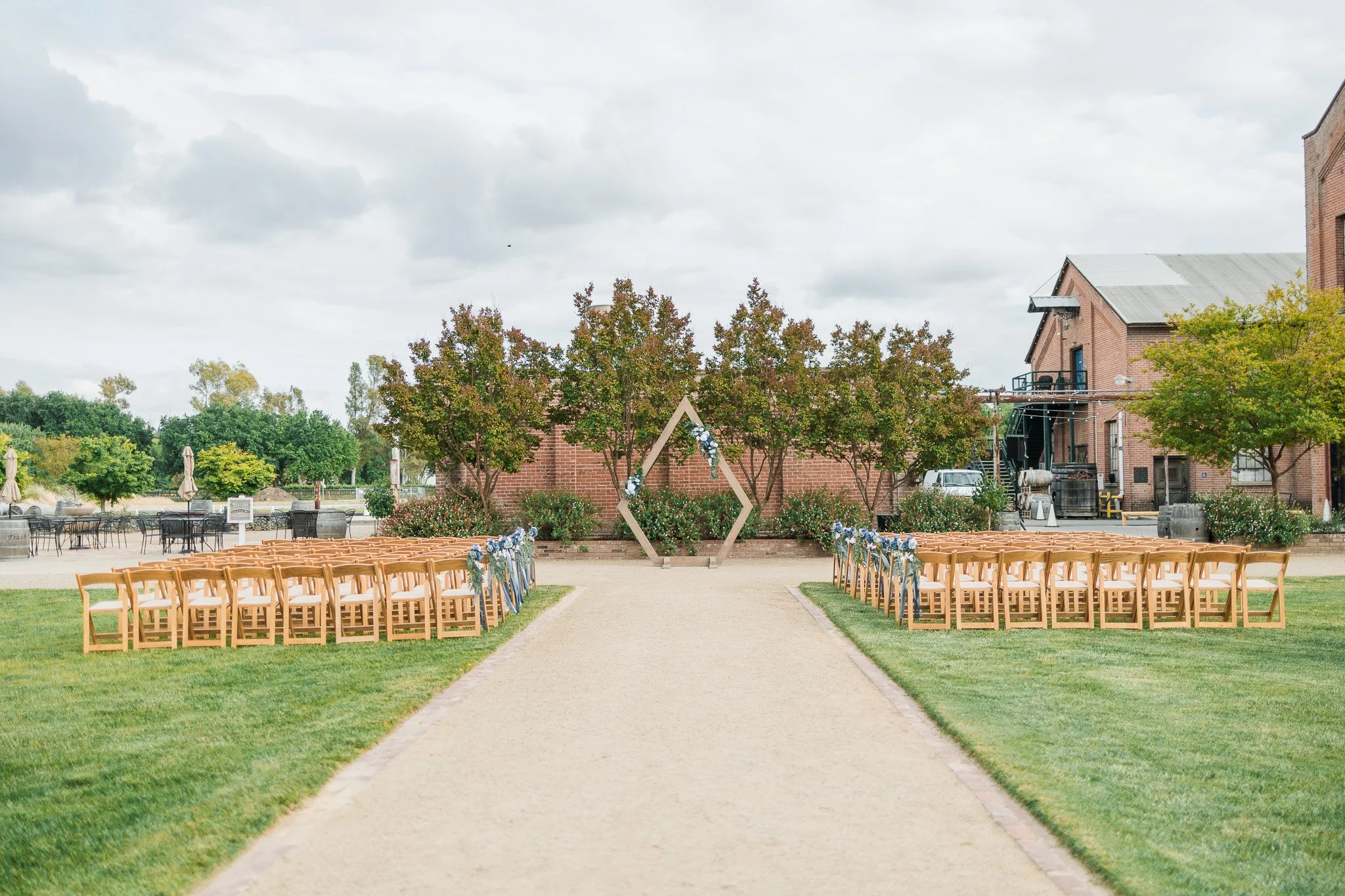 Outdoor wedding ceremony setup with rows of wooden chairs decorated with blue and white flowers, a geometric wooden arch with floral arrangements, on a gravel pathway surrounded by green grass and trees, with brick buildings and an overcast sky in th