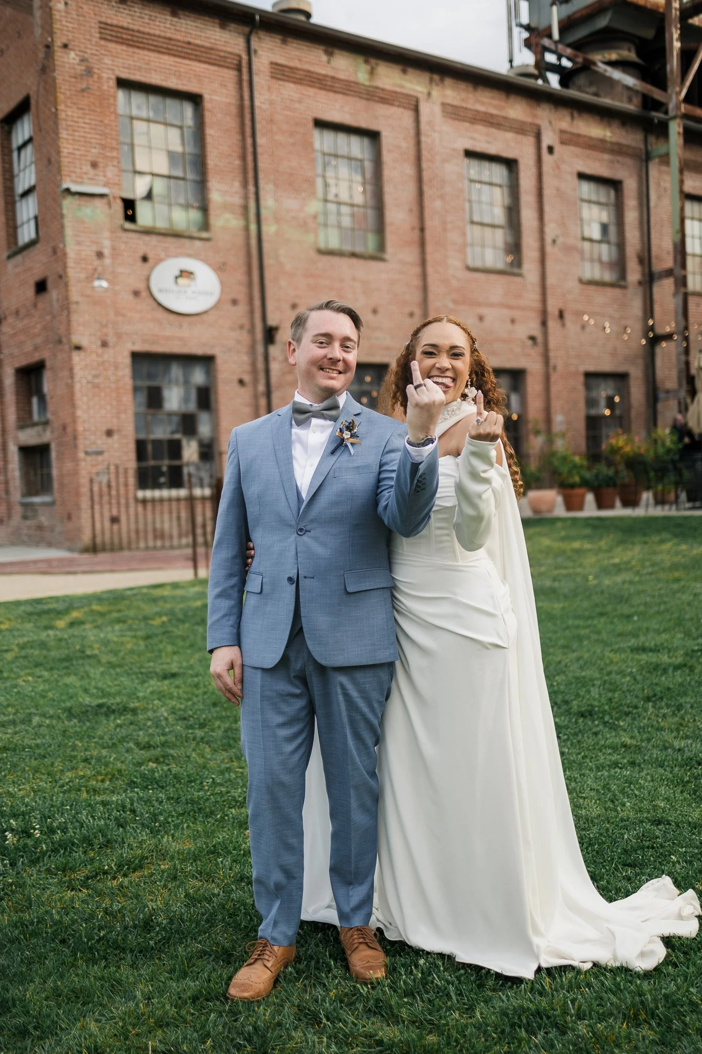 A happy couple in wedding attire standing outside in front of a brick building, making rude hand gestures and smiling.