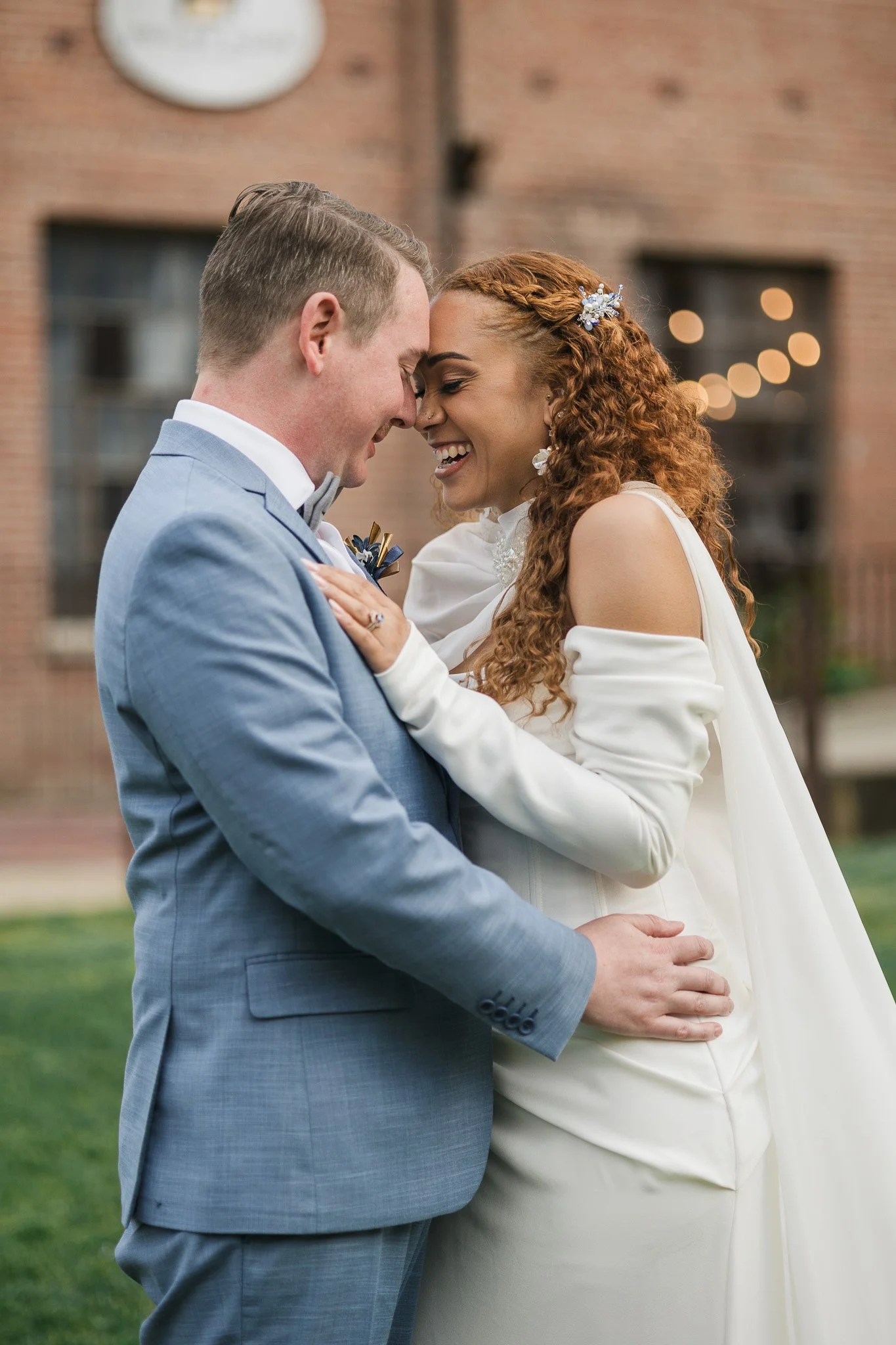 A newly married couple standing close together outdoors, smiling with foreheads touching, with a brick building in the background.