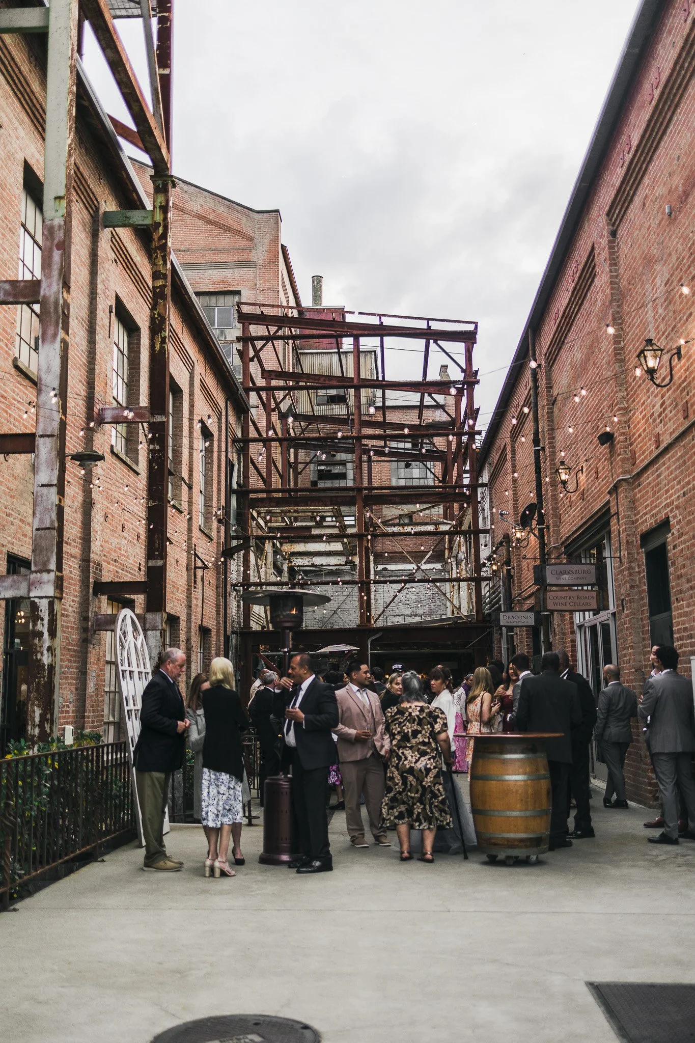 People gathered in an outdoor courtyard between brick buildings, with string lights hanging overhead and metal scaffolding structure in the background.