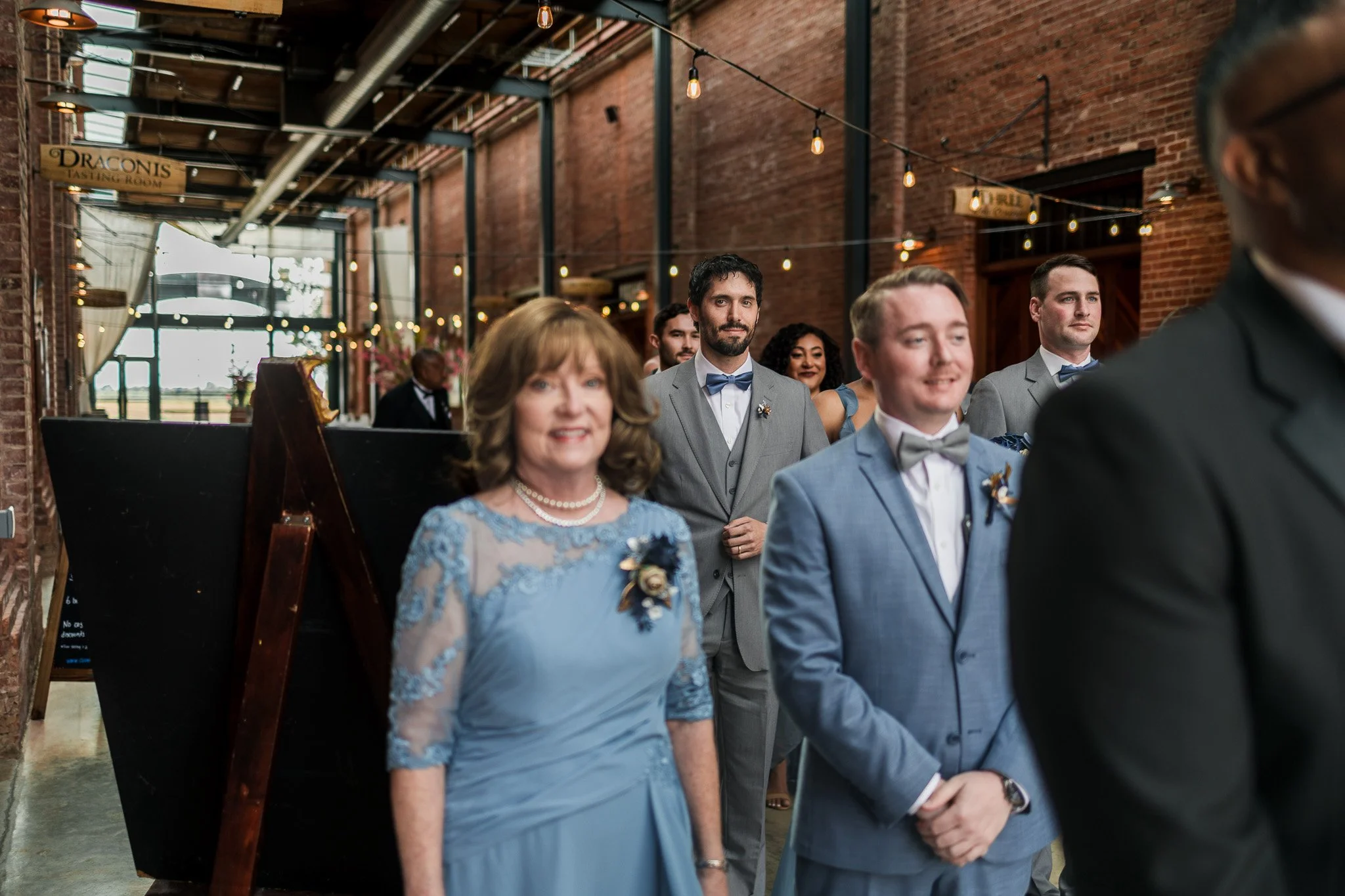 Group of formally dressed people at a wedding ceremony inside a brick-walled venue with string lights.