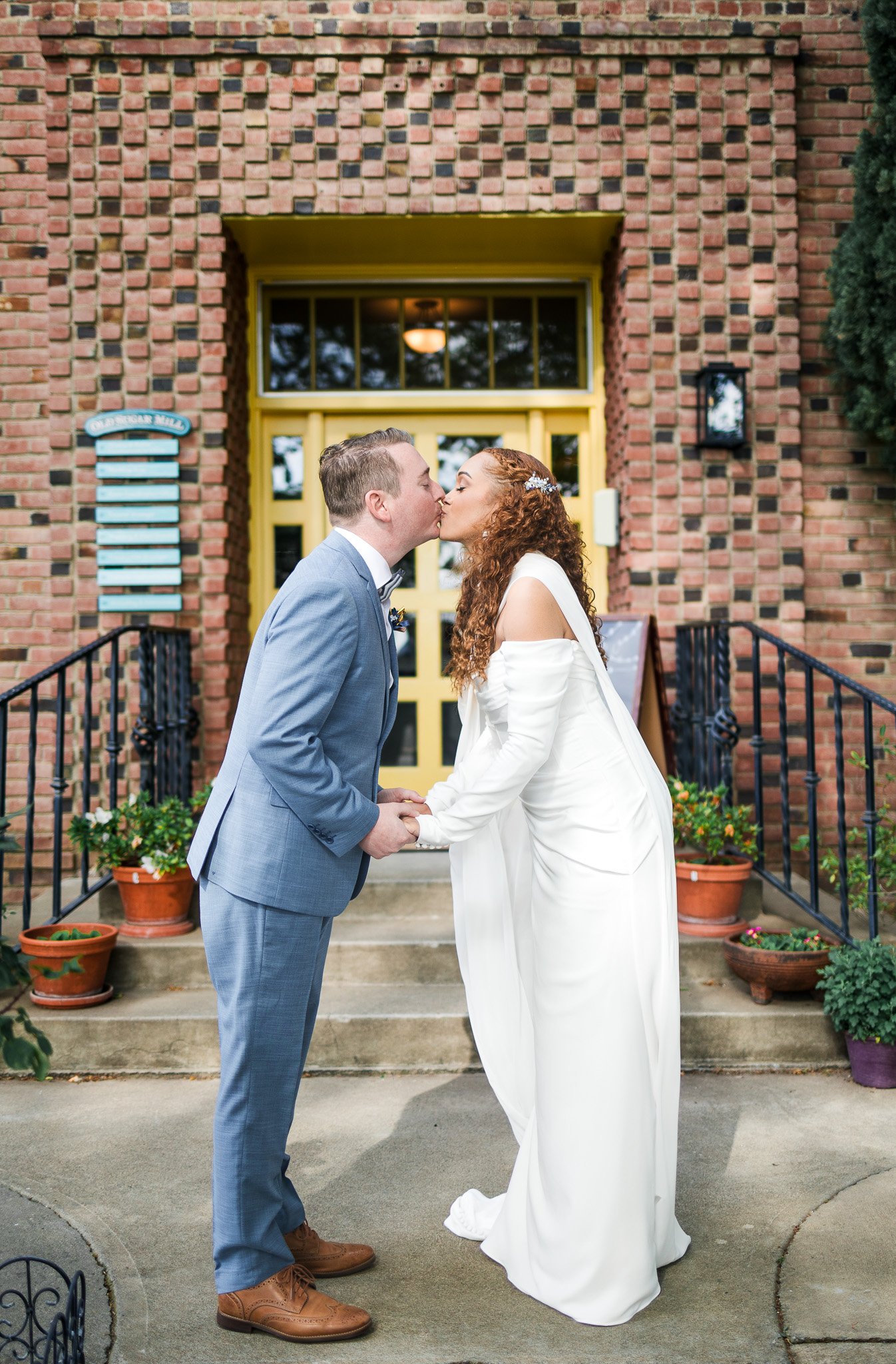 A newlywed couple kisses in front of a brick building with a yellow door, holding hands and dressed in wedding attire.