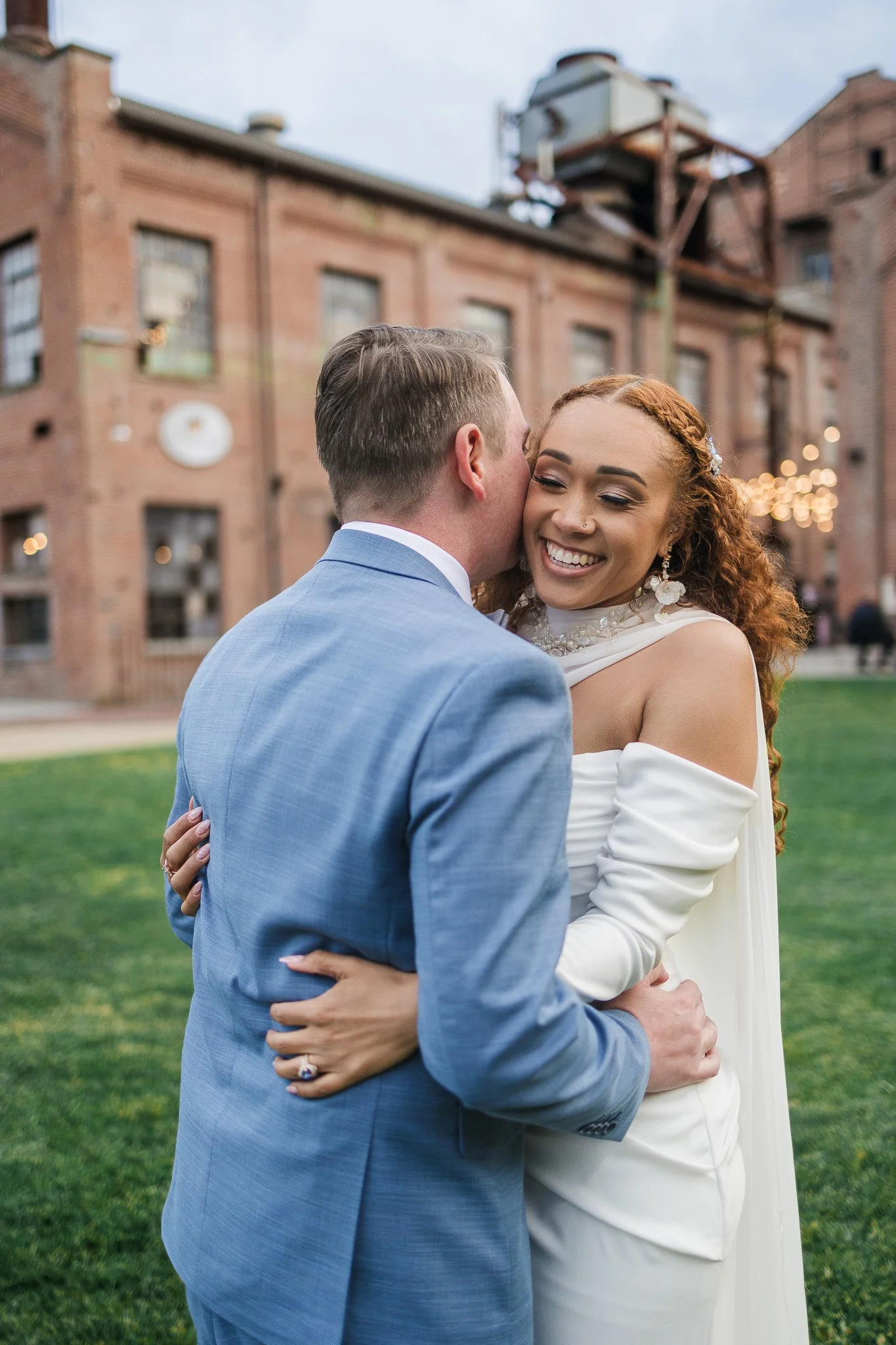 A smiling bride with red curly hair and a groom in a blue suit sharing a kiss outdoors on a grassy area with a brick building in the background.