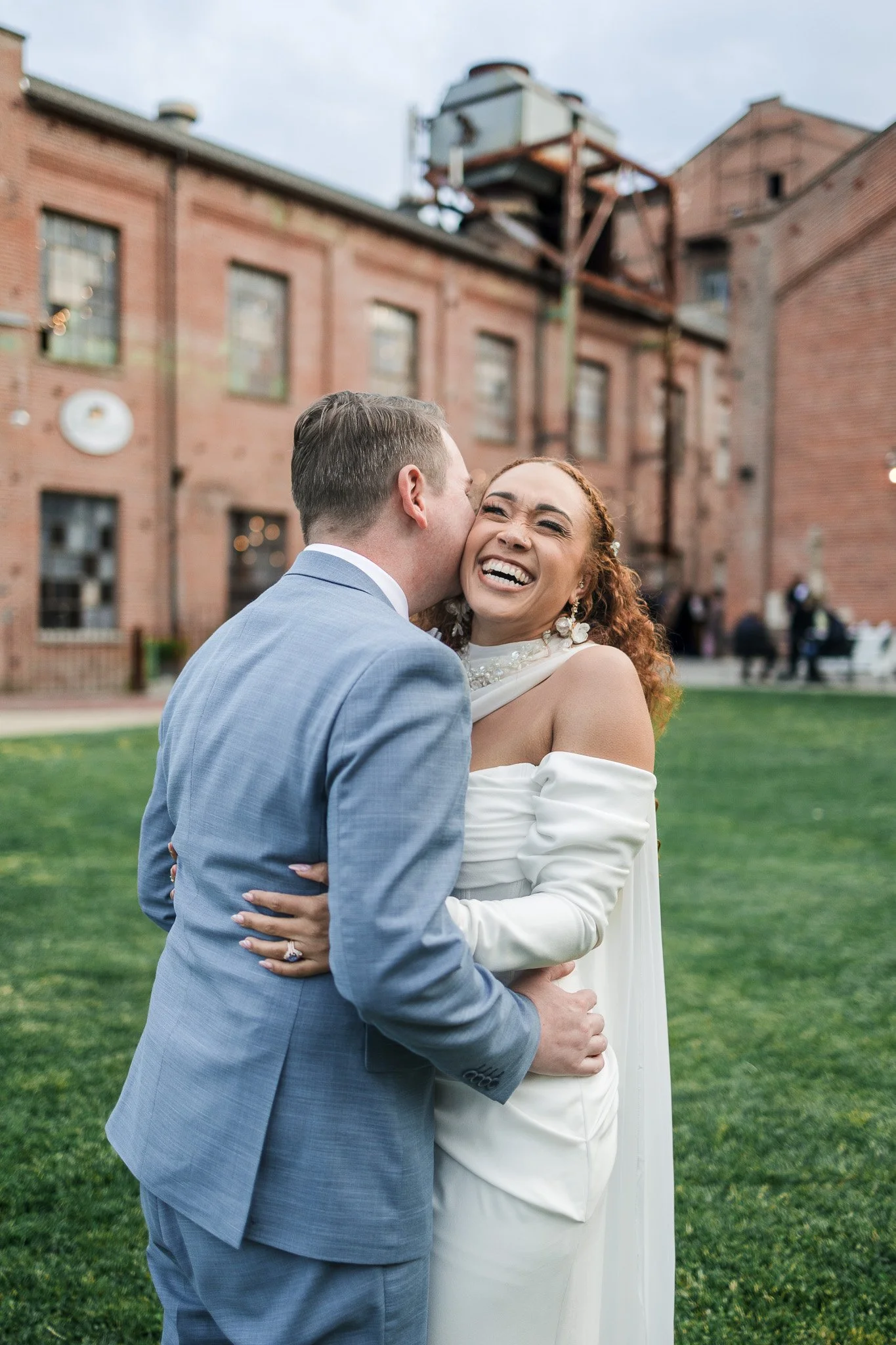 A happily smiling bride and groom hug outdoors near a red brick building, with the groom kissing the bride on the cheek.