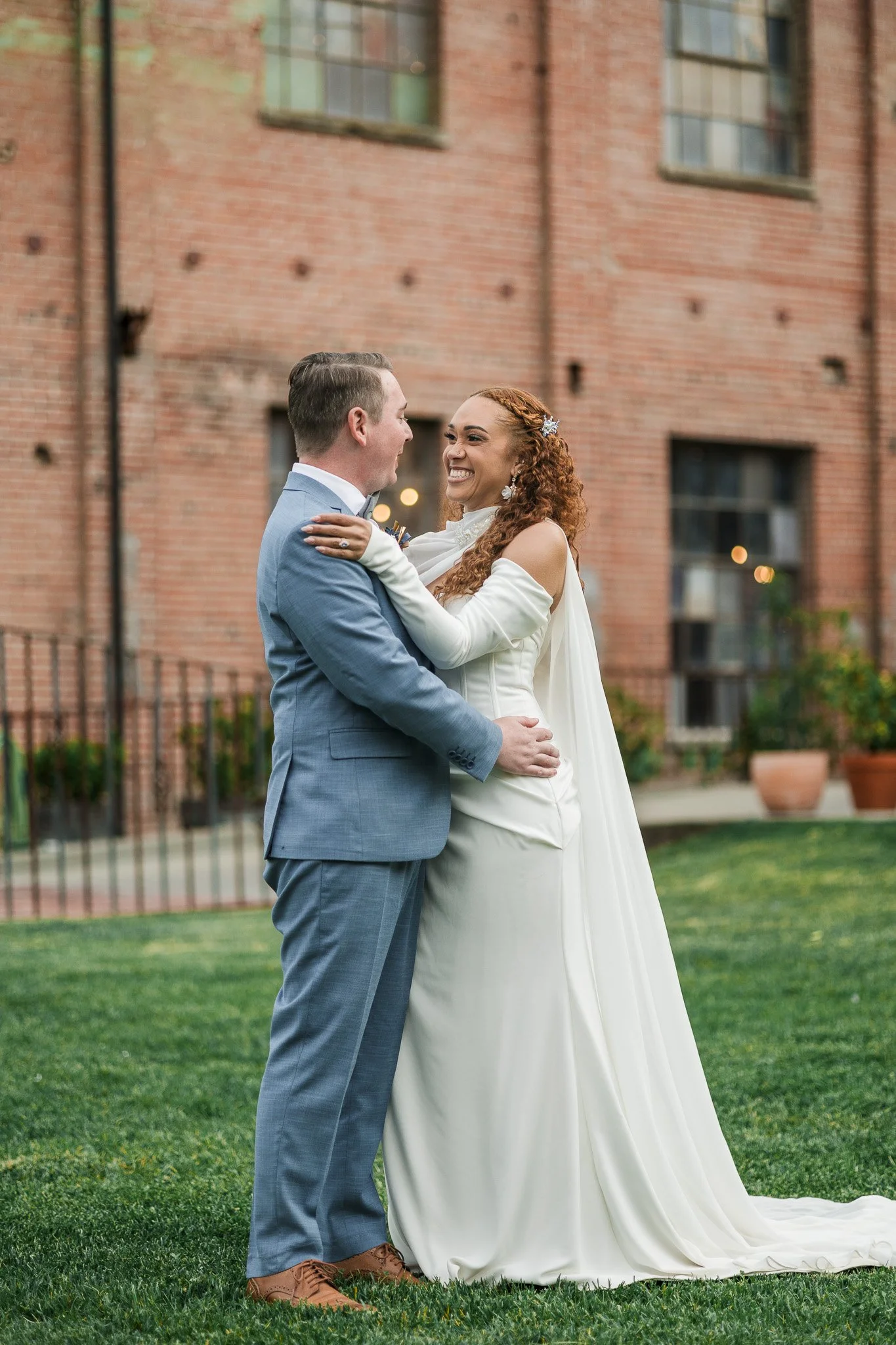 A bride and groom happily embrace outdoors in front of a brick building during their wedding.