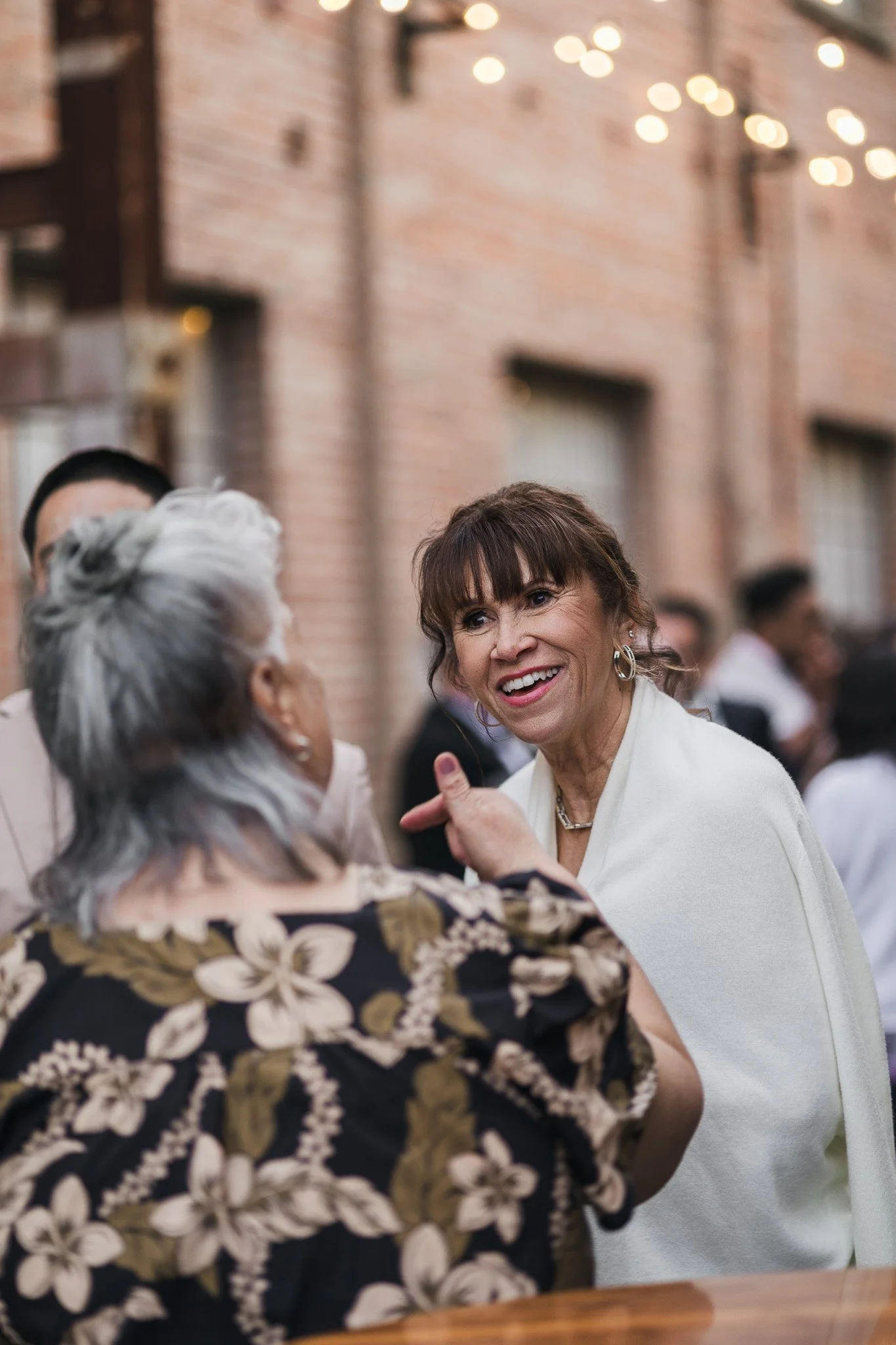 Two women engaged in conversation at a social gathering outdoors, with a brick building background and string lights overhead.